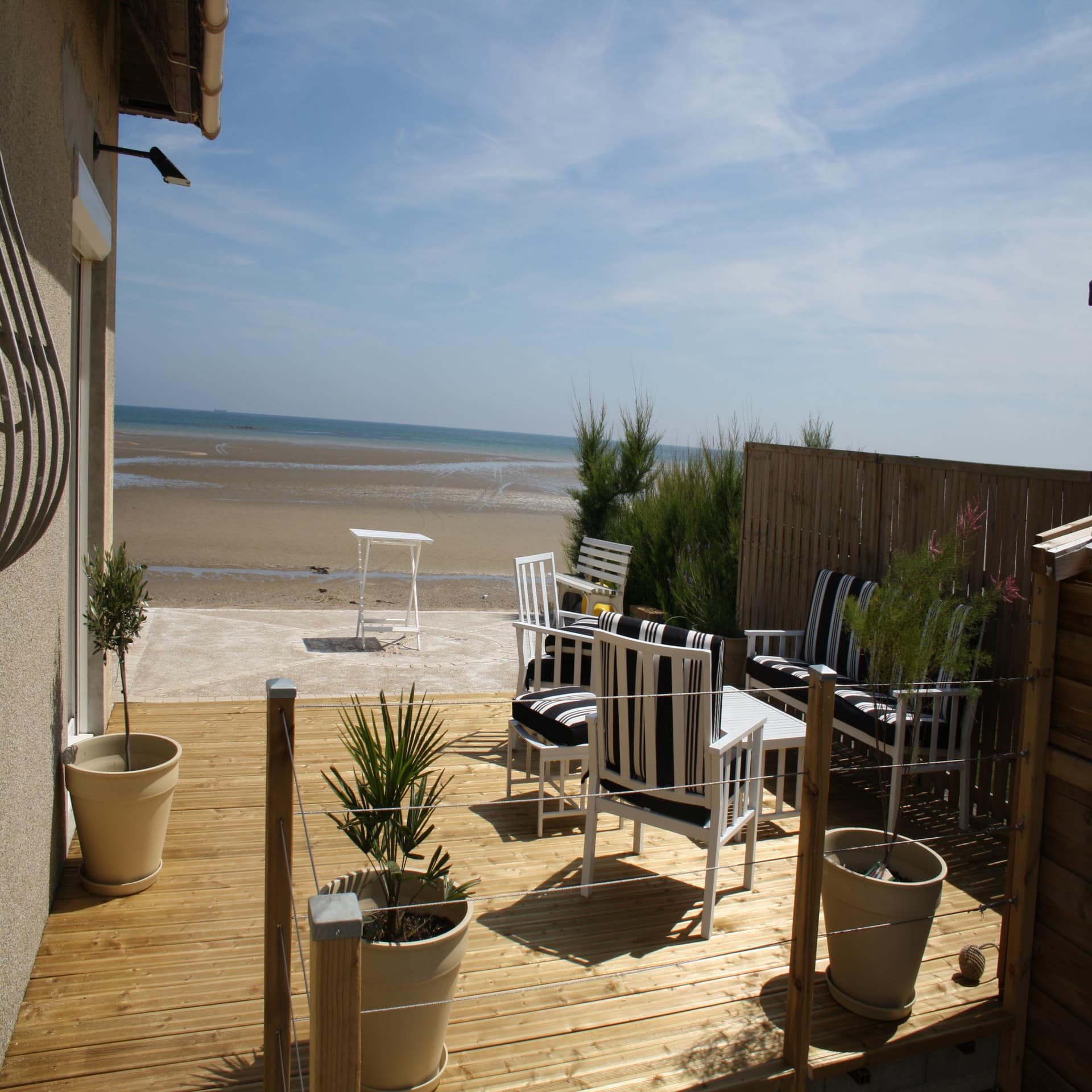 Holzterrasse mit Sitzgelegenheiten und Pflanzenkübeln von einem Haus direkt am Strand mit Blick auf das Meer. 