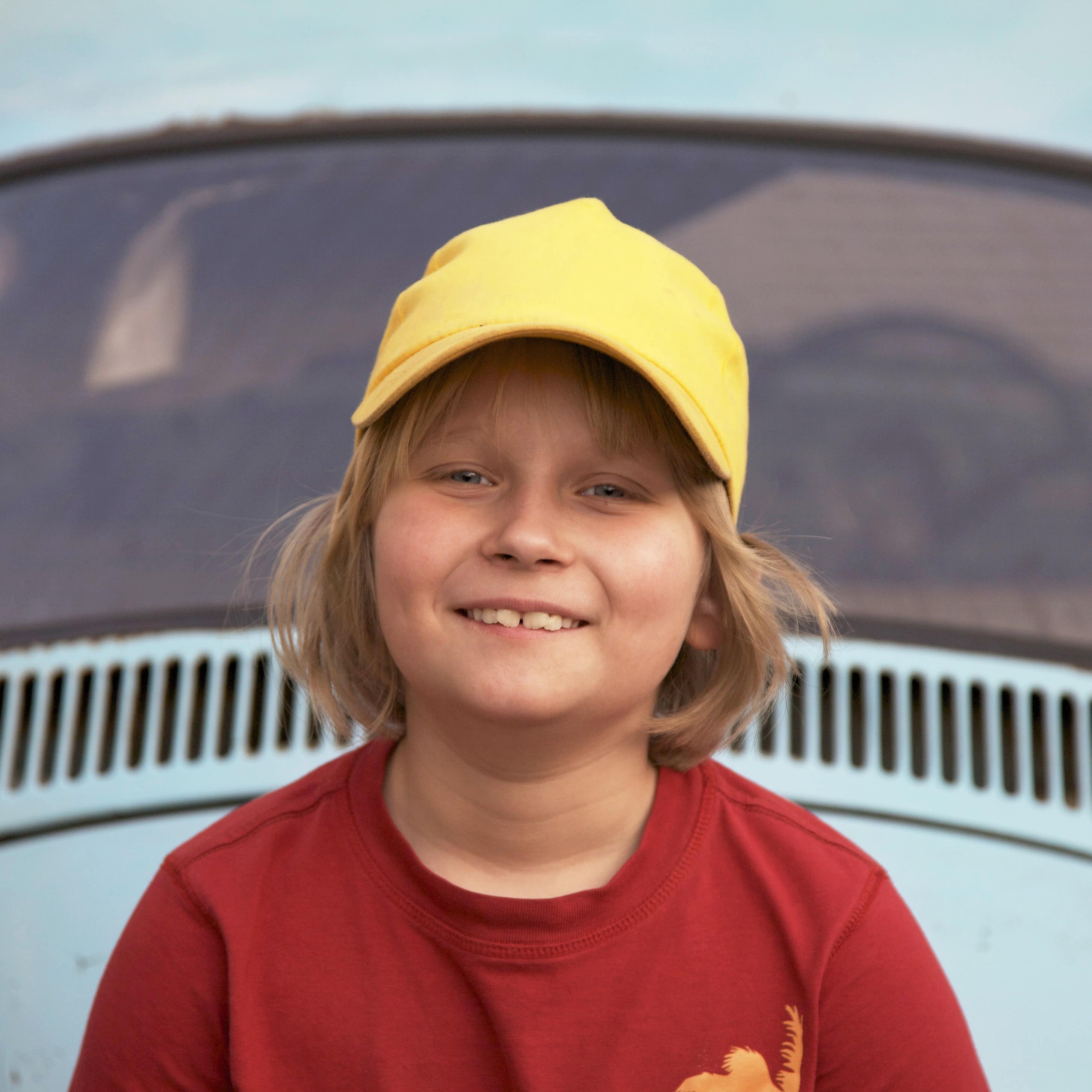 Nahaufnahme: Ein June in rotem T-Shirt und mit gelber Mütze vor einem VW Käfer.