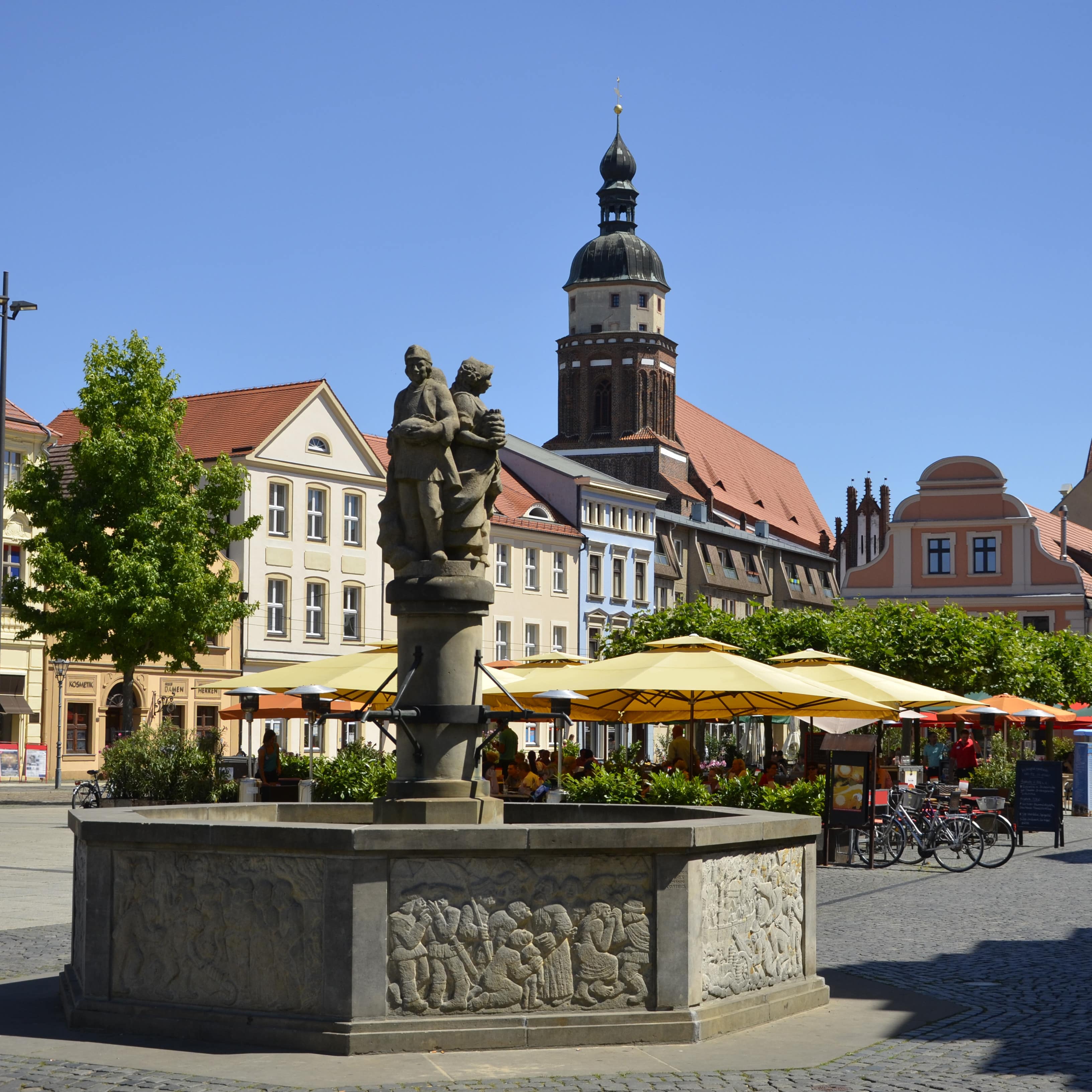 Blick auf den Marktbrunnen am Altmarkt, dahinter ein Café, Häuser und eine Kirche.