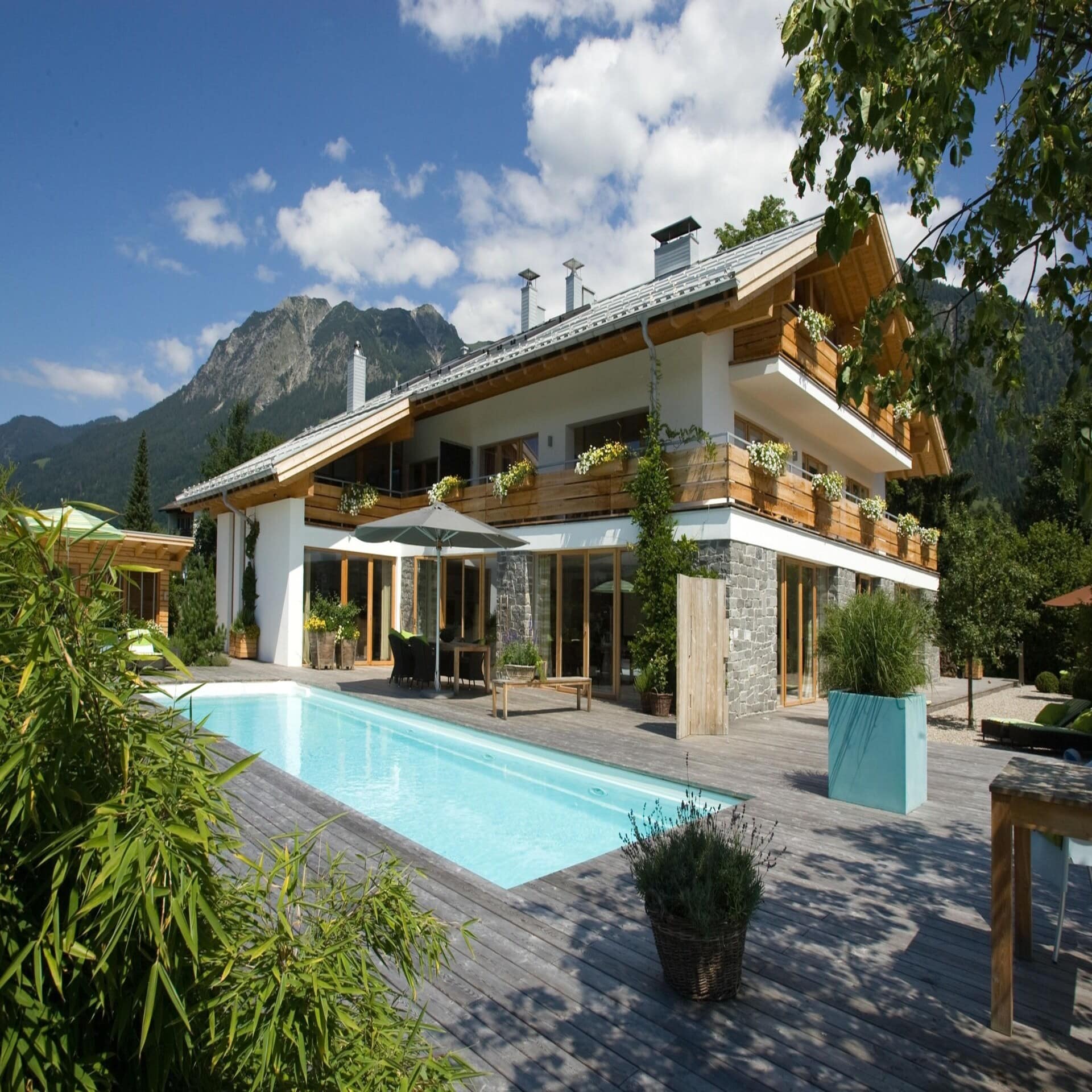 Ferienvilla im bayerischen Stil mit Holzdeck und großem Pool in Oberstdorf. Die Sonne scheint.