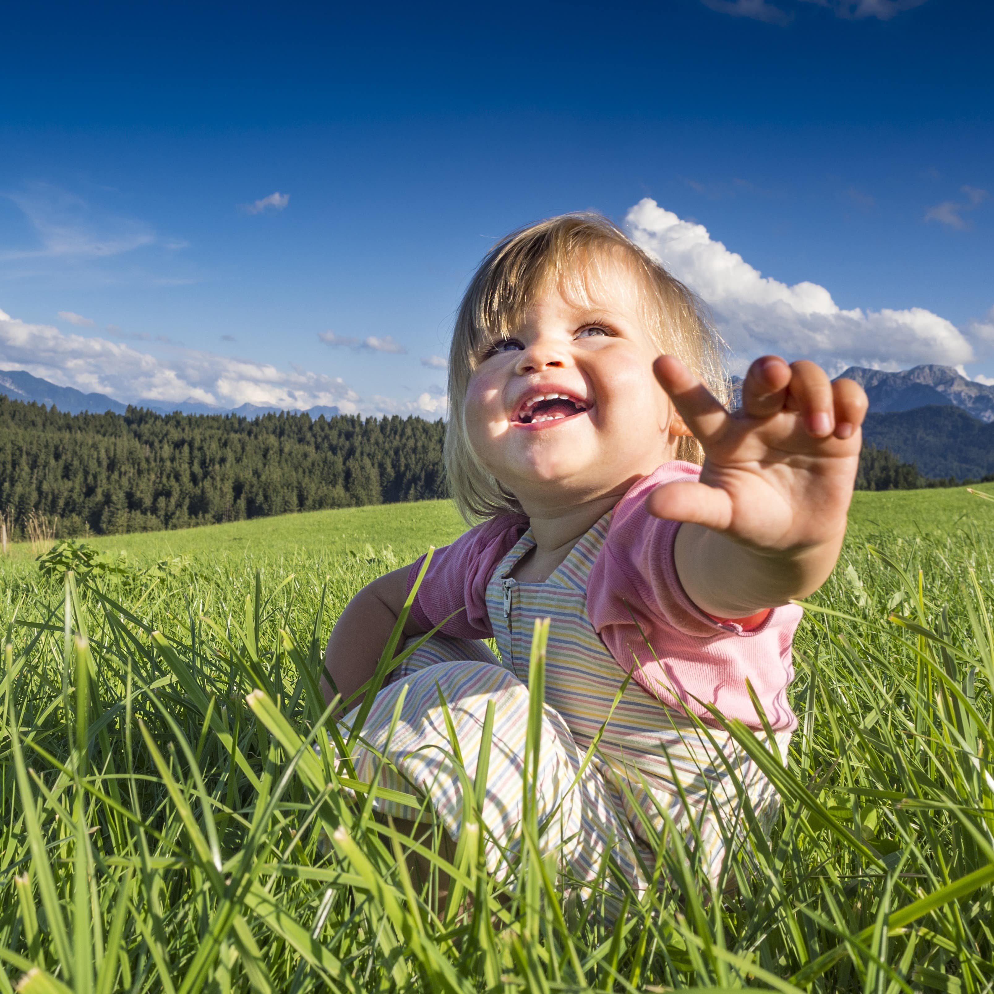 Nahaufnahme: Ein kleines Mädchen sitzt auf einer Wiese im Allgäu, im Hintergrund Wald und Berge.