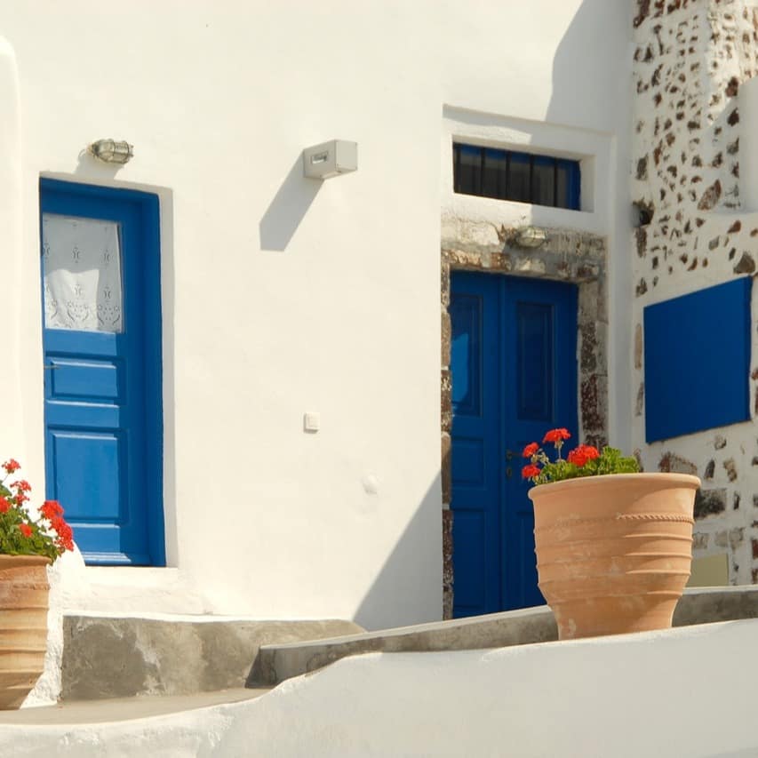 Ein Haus in landestypischer Architektur und blau weißer Fassade, davor stehen Töpfe mit roten Blumen.