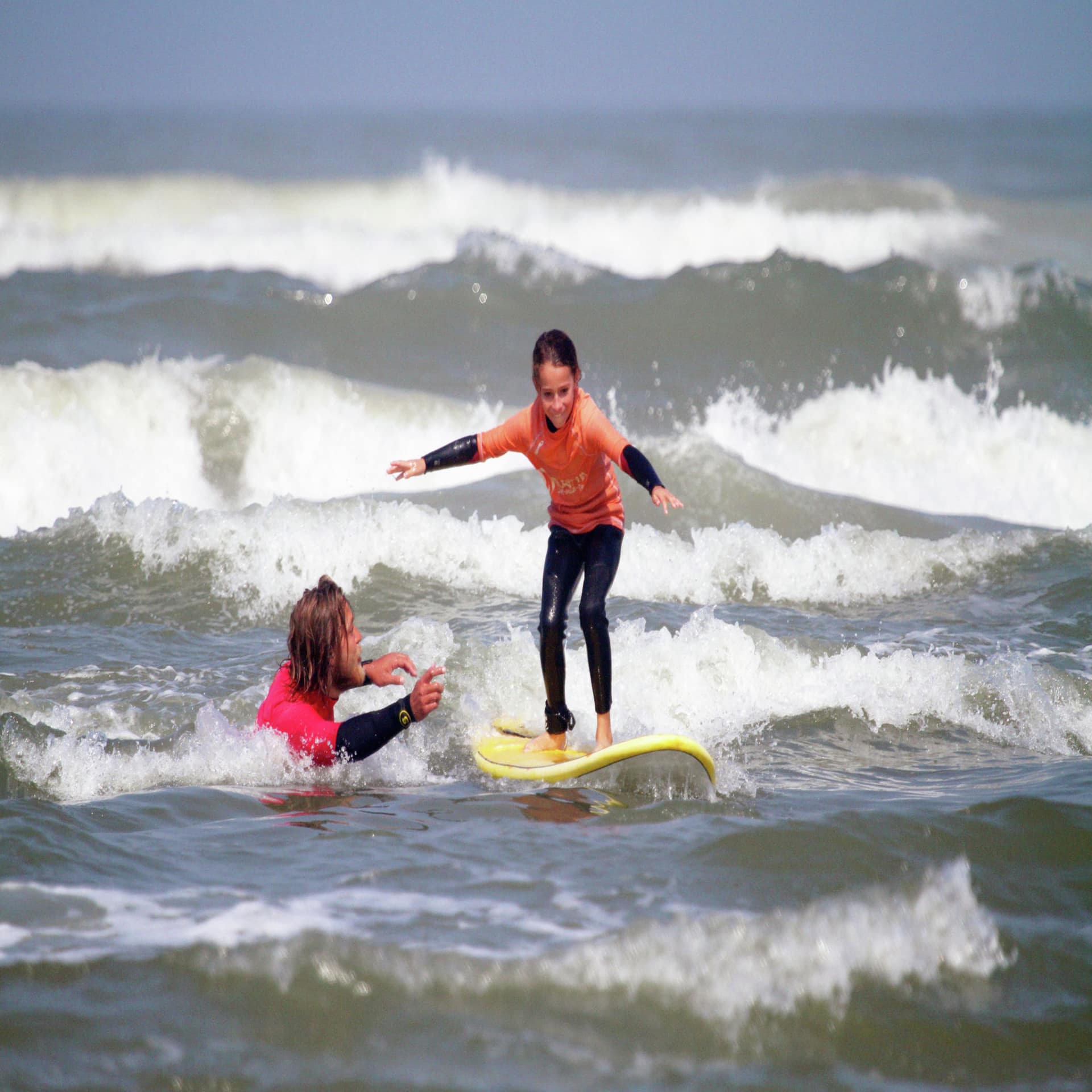 Kind im Wetsuit auf einem Surfbrett im Wasser, ein Mann gibt Surfunterricht. 