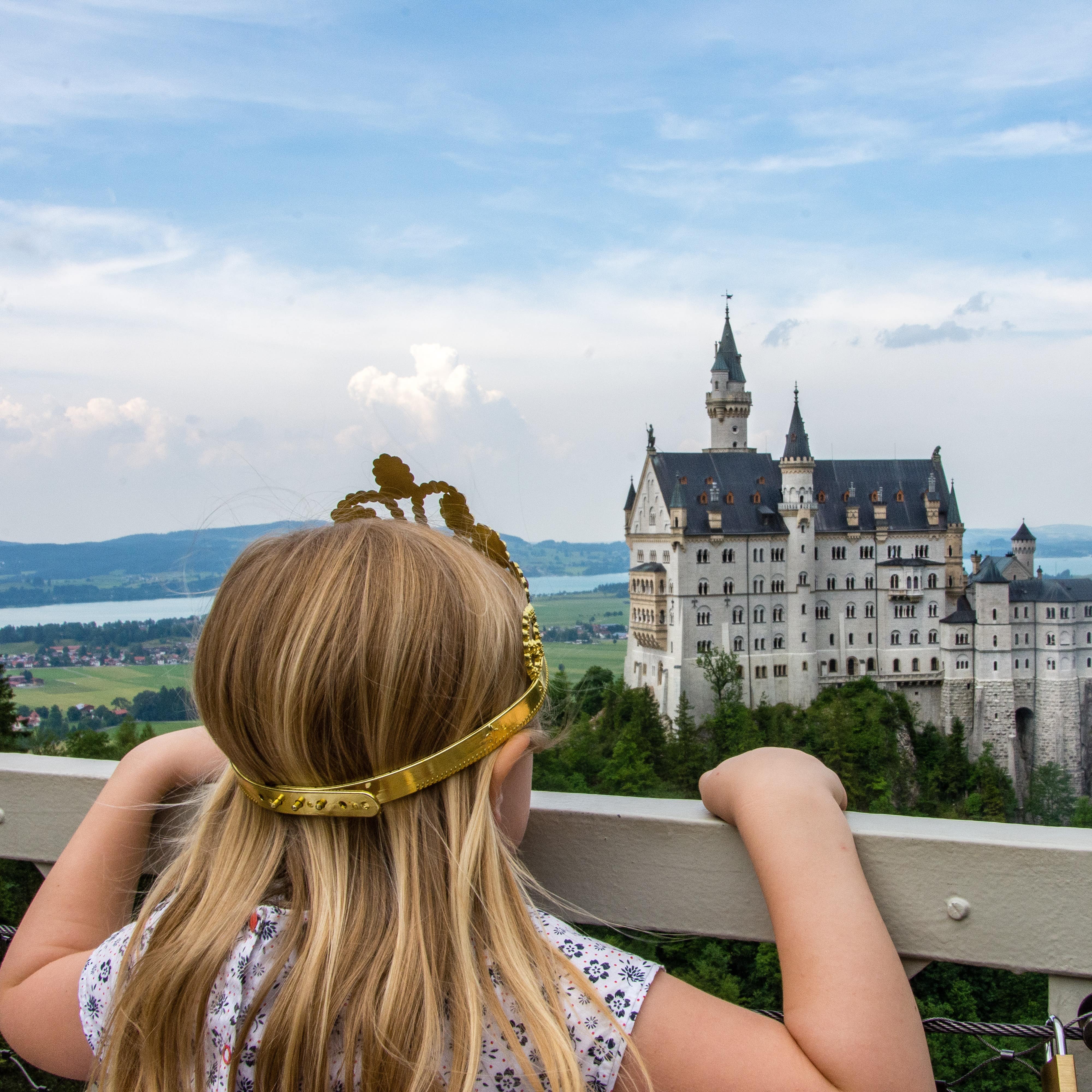 Rückenansicht: Ein kleines Mädchen mit langen blonden Haaren und eine Krone auf dem Kopf schaut auf das Schloss.