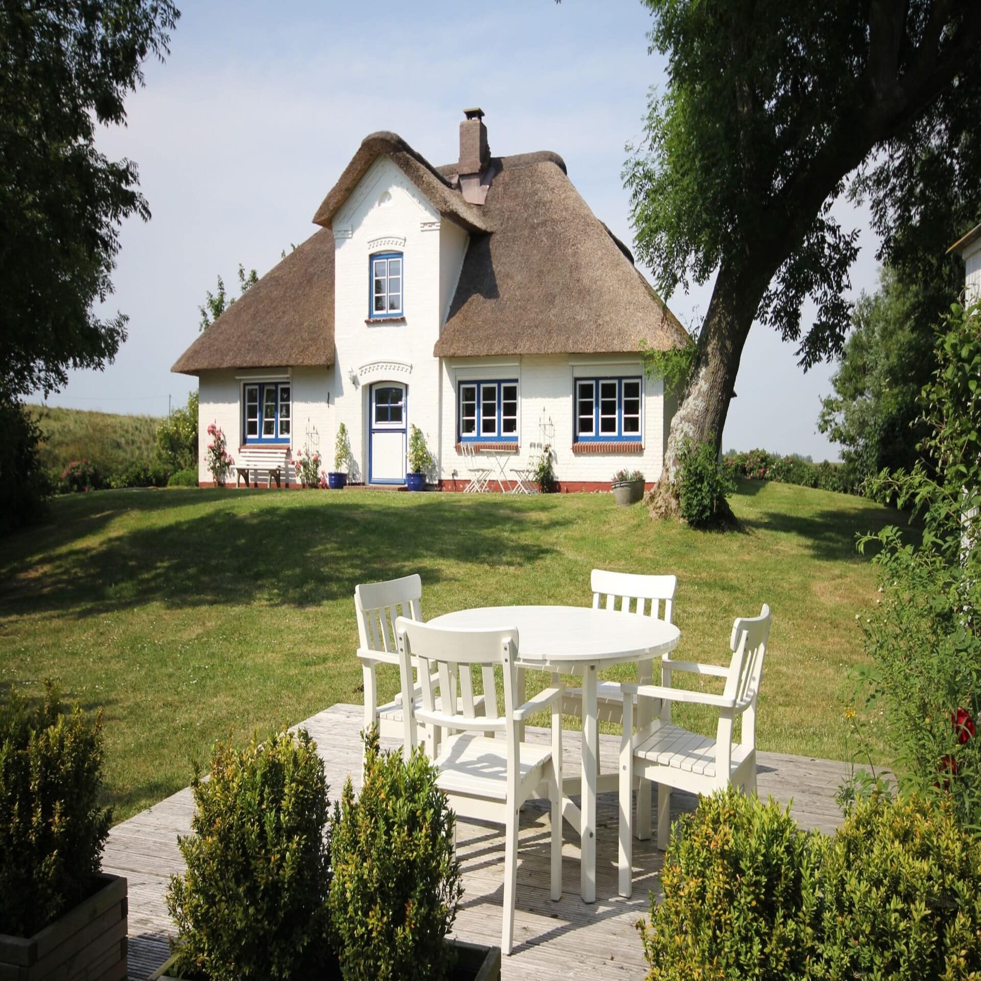 Blick von einer Terrasse mit weißen Möbeln auf ein Friesenhaus mit Reetdach in einem Garten.
