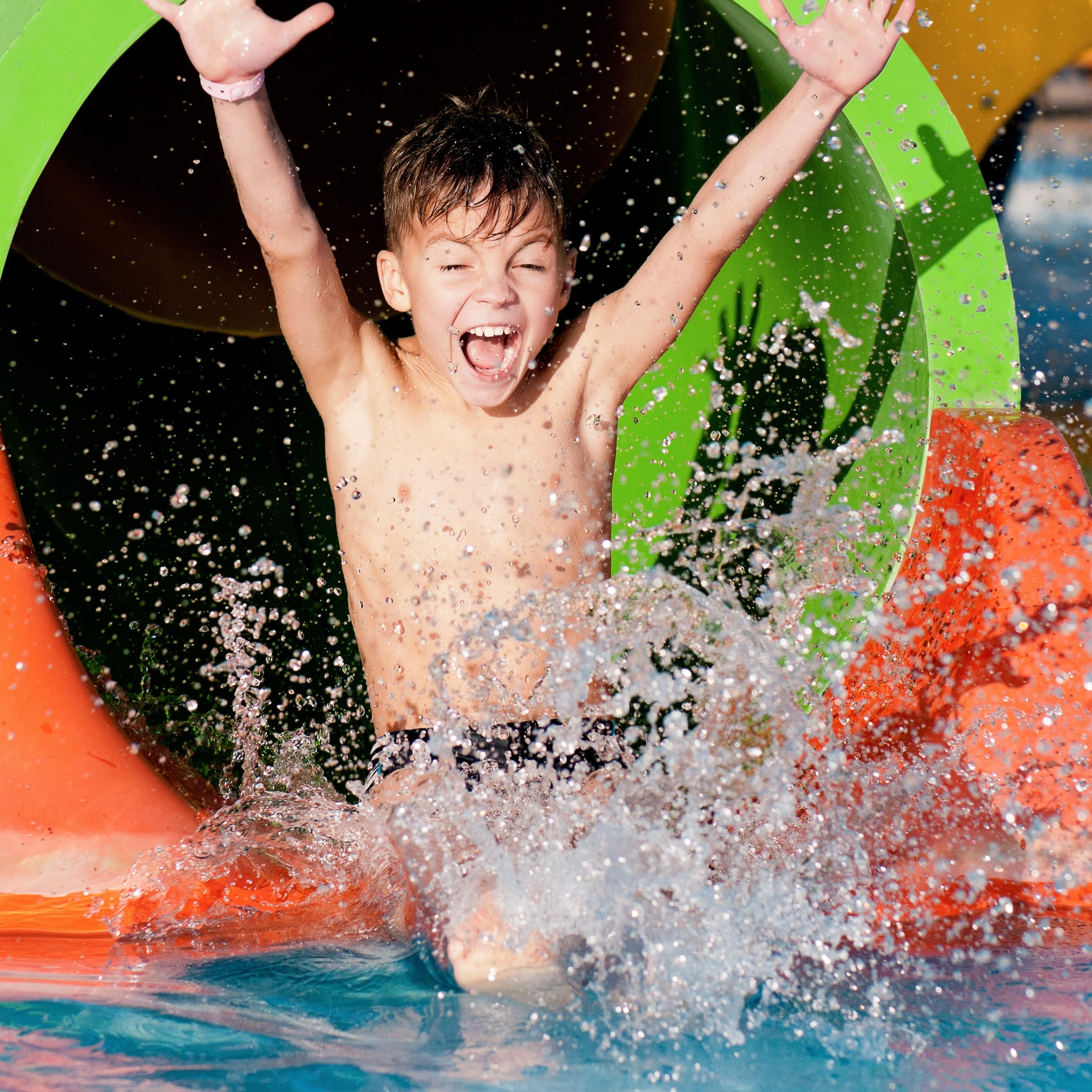 Ein Junge saust aus einer Wasserrutsche ins Wasser und reißt die Arme hoch.
