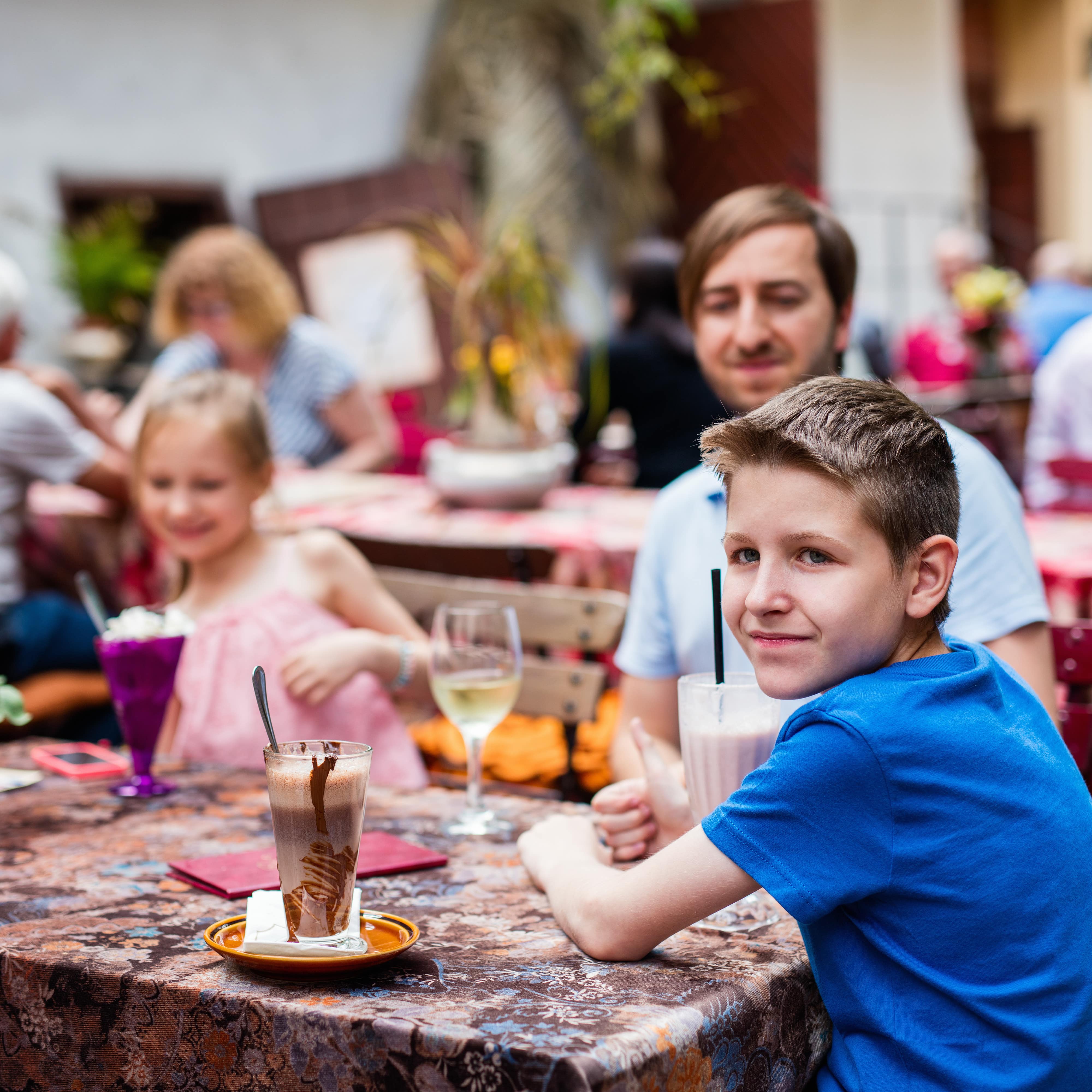 Vater und 2 Kinder an einem Tisch draußen in einem Restaurant, die Kinder trinken Milchshake, der Vater Wein.