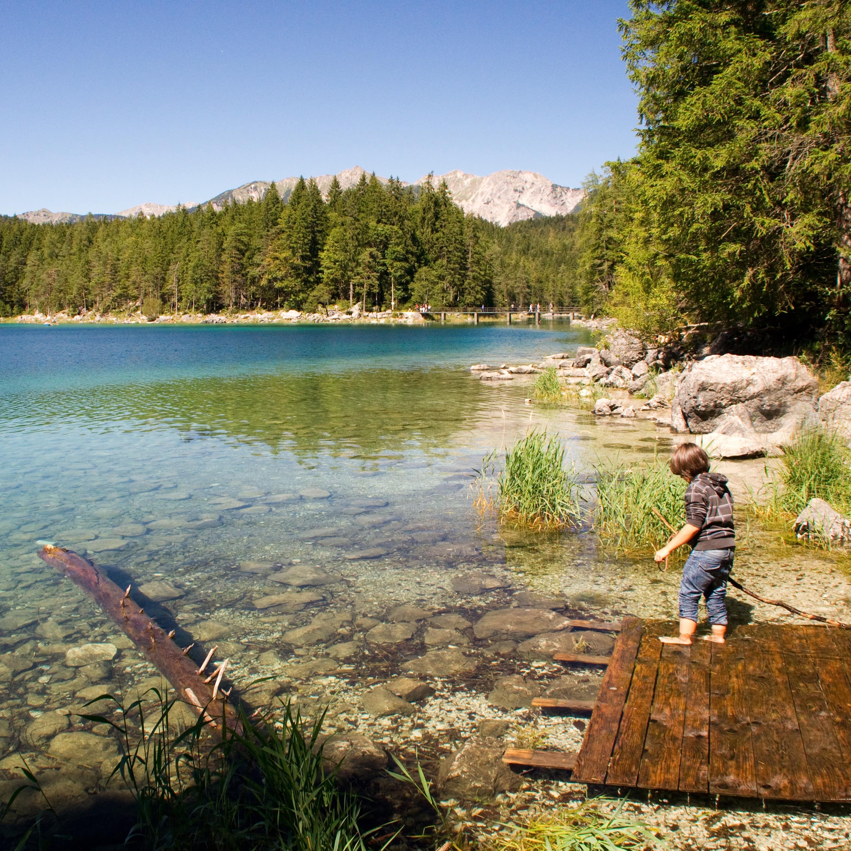 Ein kleiner Junge auf Holzplanken und mit einem Stock in der Hand am Wasser des Eibsee.