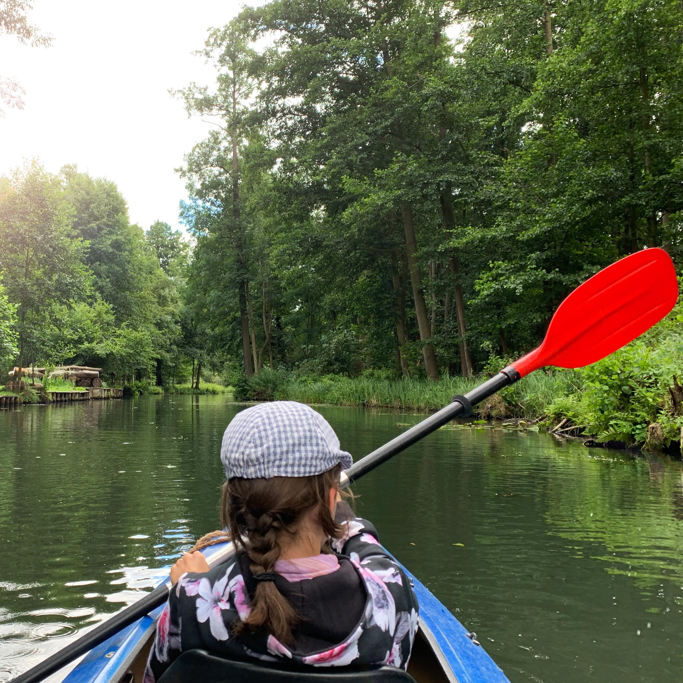 Rückenansicht: Ein Mädchen mit Zopf und karierter Mütze rudert in einem Kanu durchs Wasser. Links und rechts Bäume.