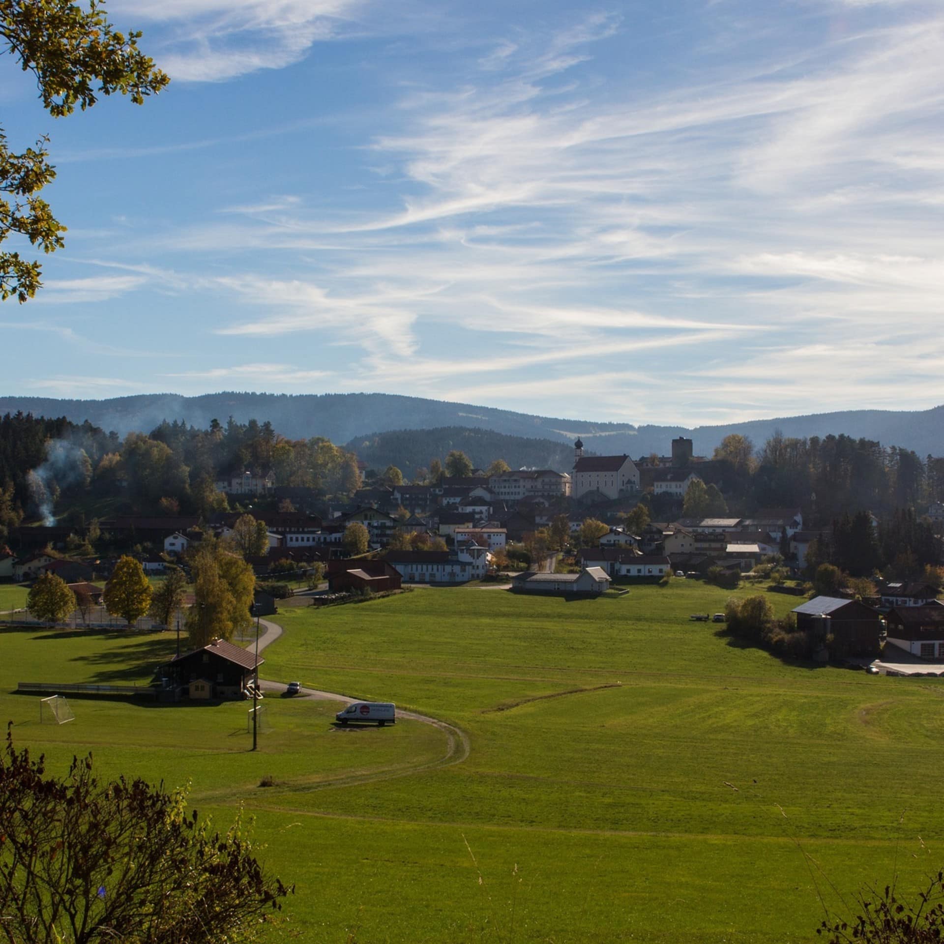 Panoramablick auf ein Dorf im Bayerischen Wald, im Vordergrund ist eine grüne Wiese zu sehen. 