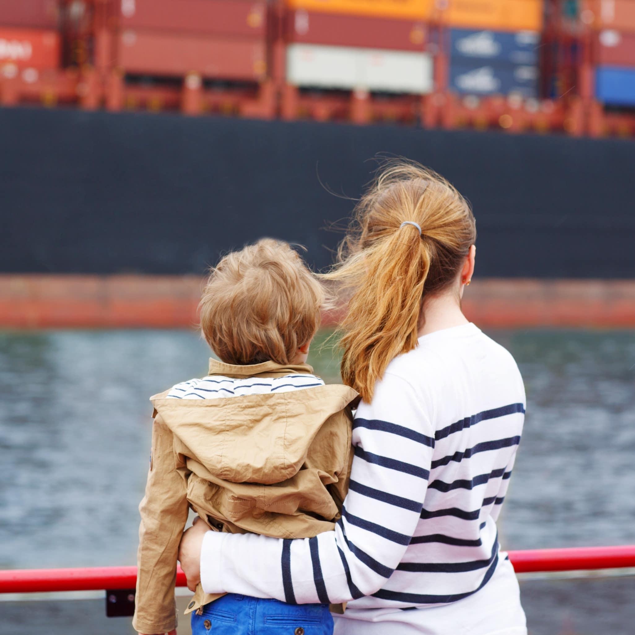 Rückenansicht: Mutter und Sohn an der roten Reling eines Boots blicken auf ein Containerschiff.