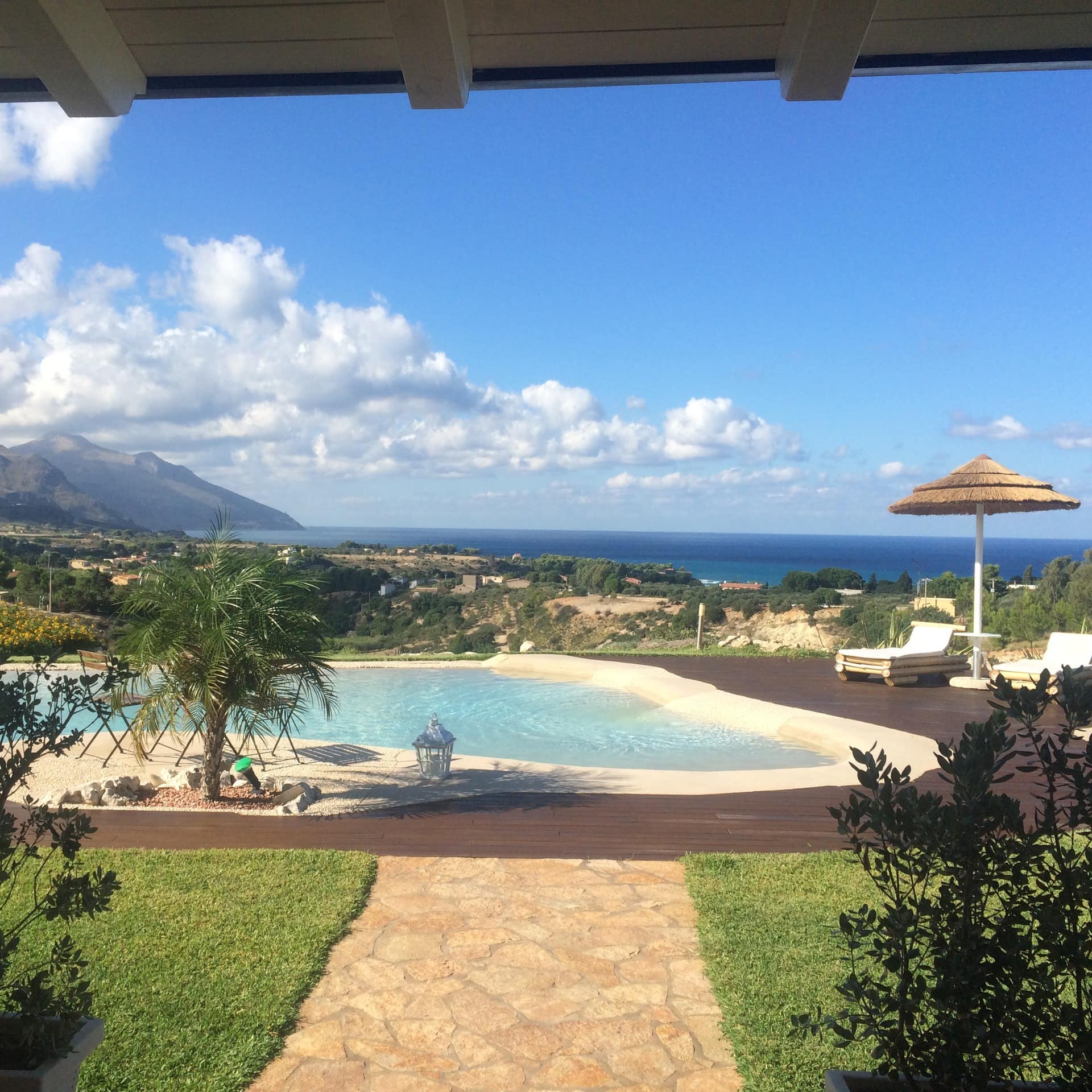 Blick von der Terrasse eines Ferienhauses am Meer auf den Garten mit Pool und Sonnenliegen, das Meer im Hintergrund