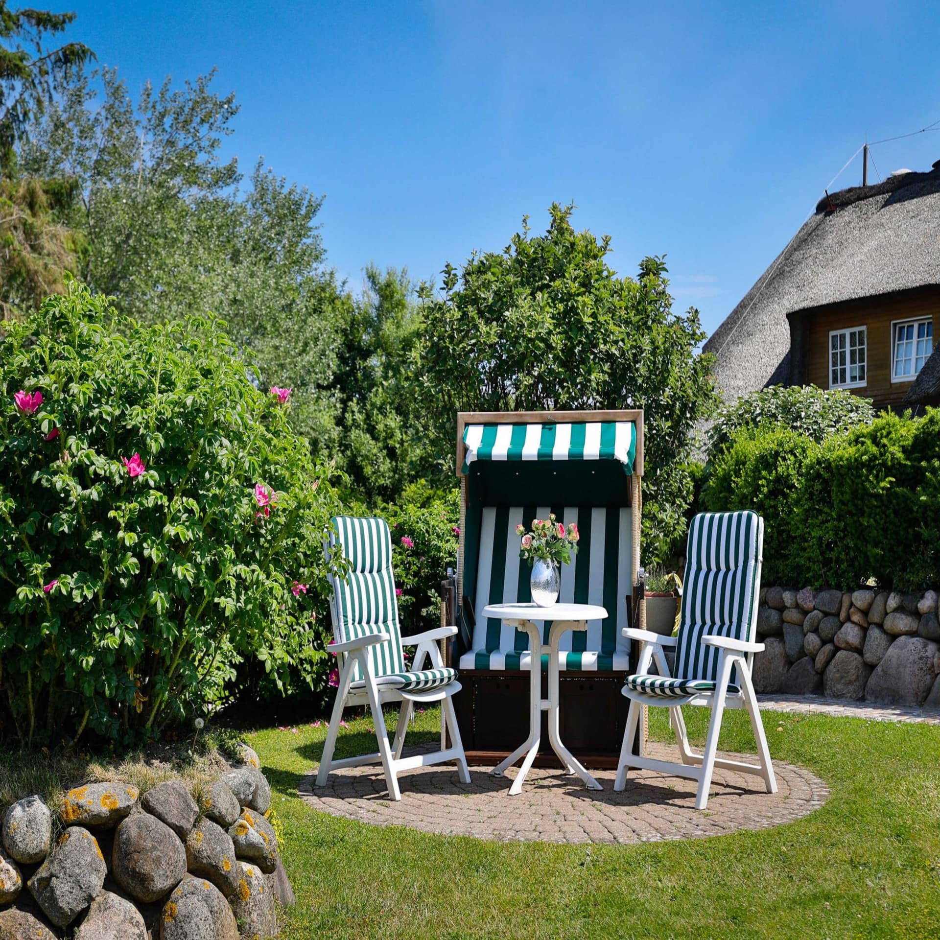 Ein grün-weiß gestreifter Strandkorb, 2 Stühle und 1 Tisch mit Blumen im Garten bei Sonnenschein.