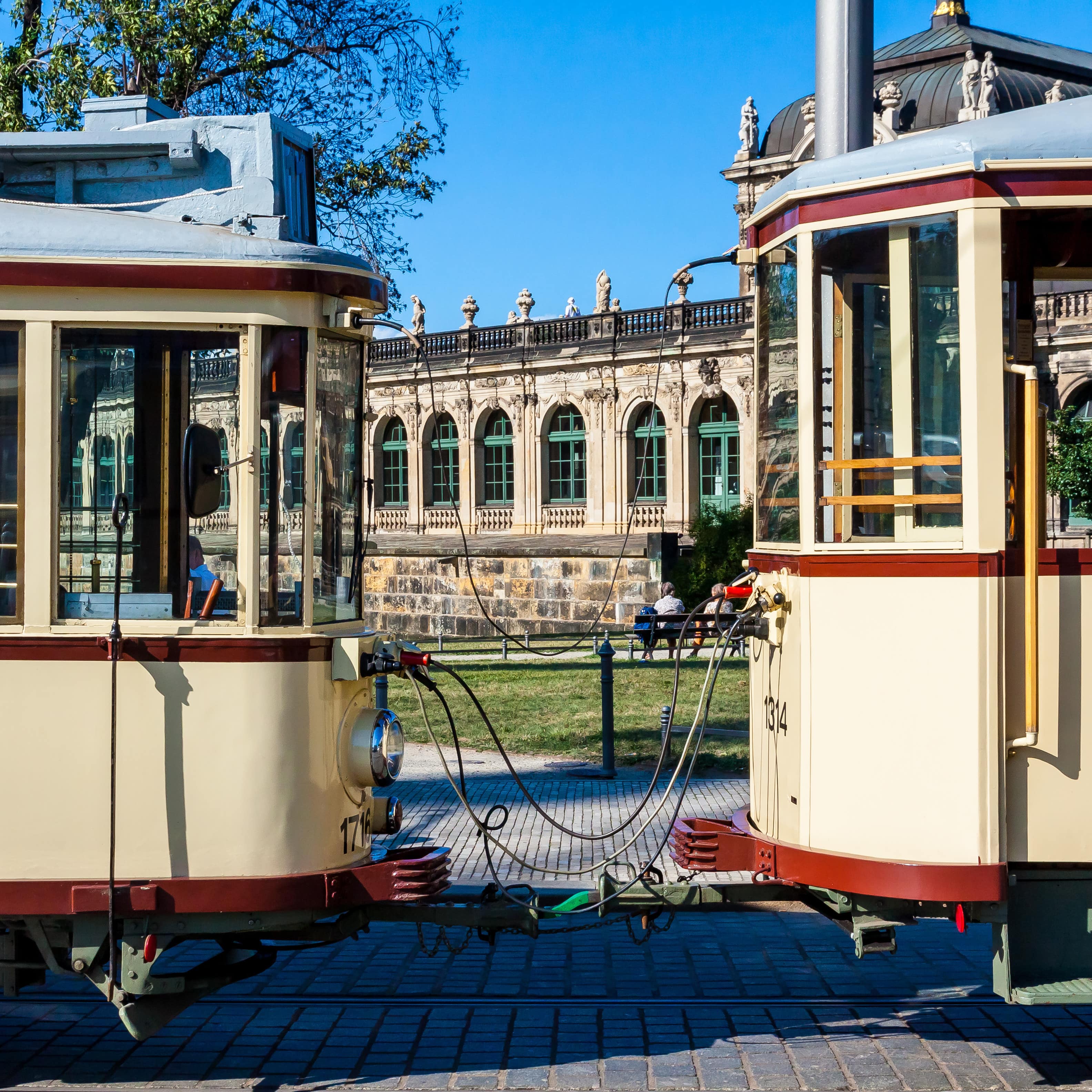 Eine historische Straßenbahn, zwischen 2 Wagen sind eine Wiese und ein Altbau zu sehen.