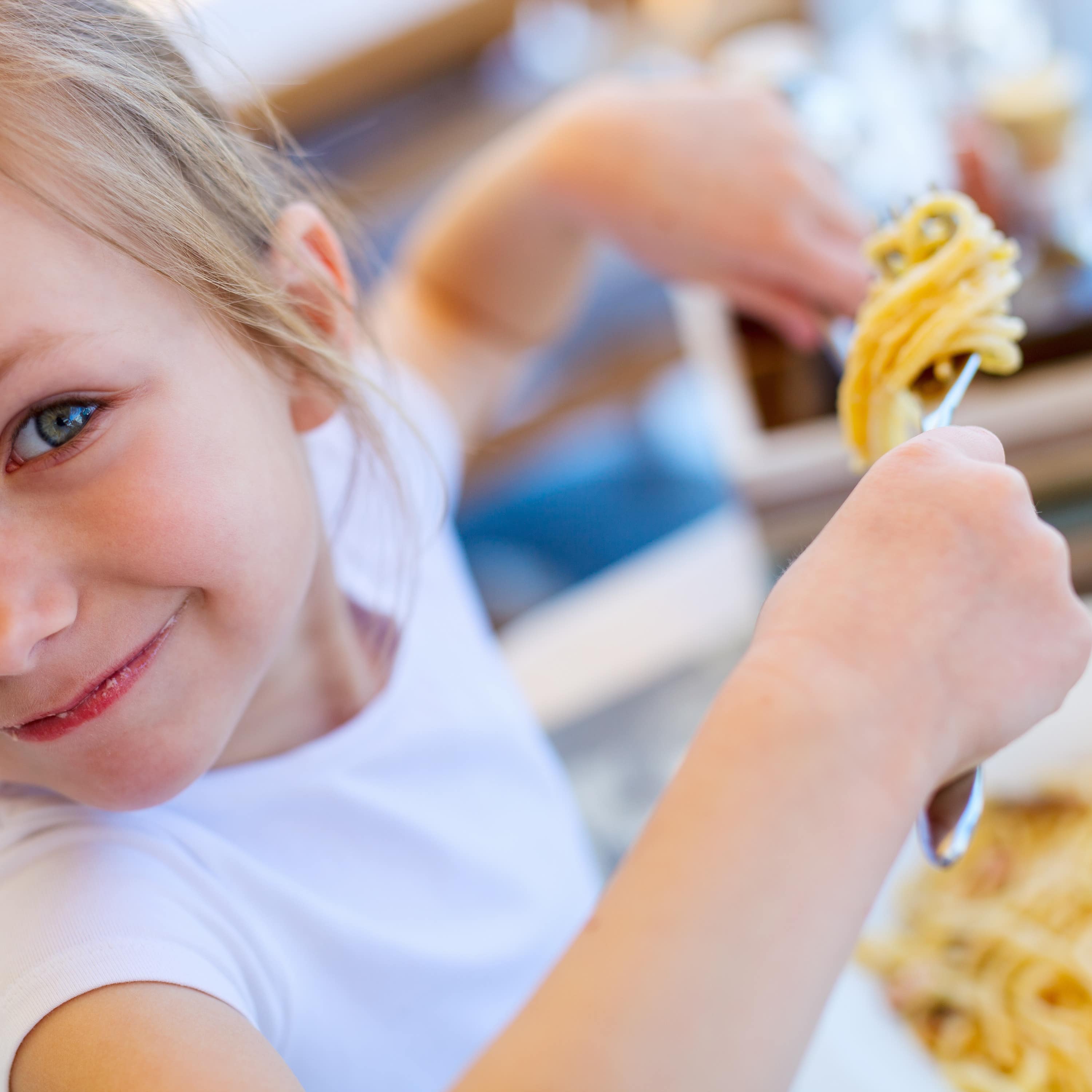 Nahaufnahme: Ein Mädchen in weißem T-Shirt isst Spaghetti und lächelt in die Kamera.
