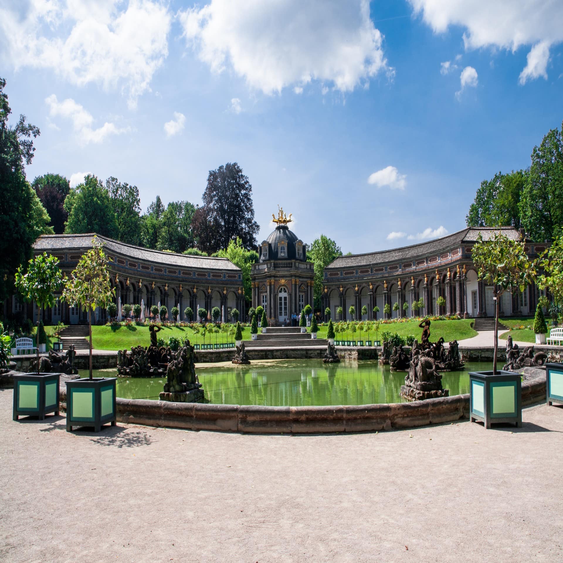 Blick auf ein Schloss in Bayreuth. Davor eine Wiese und ein Brunnen. Die Sonne scheint.