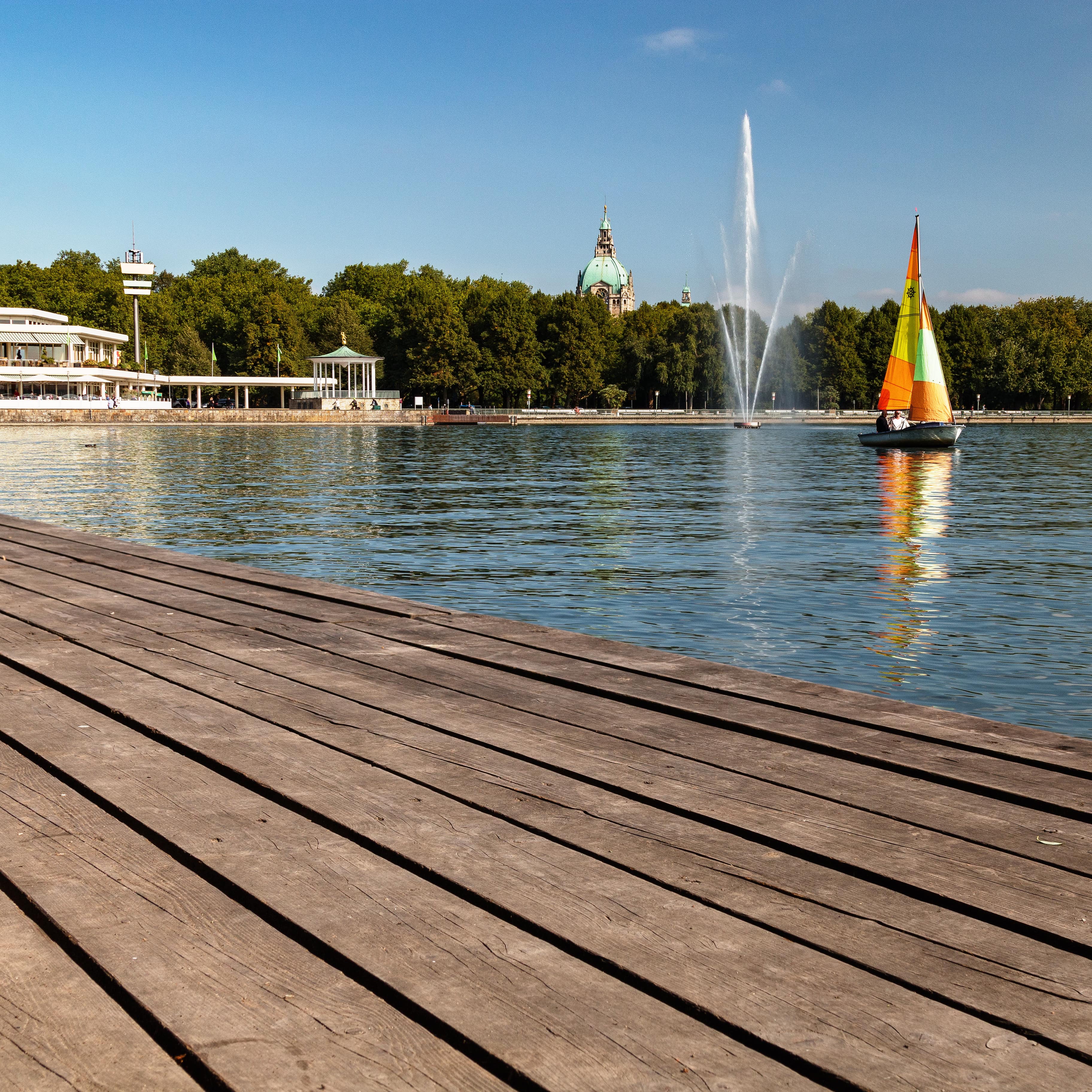 Blick über ein Holzdeck auf den Maschsee auf dem ein buntes Segelboot schippert, dahinter eine Fontäne.