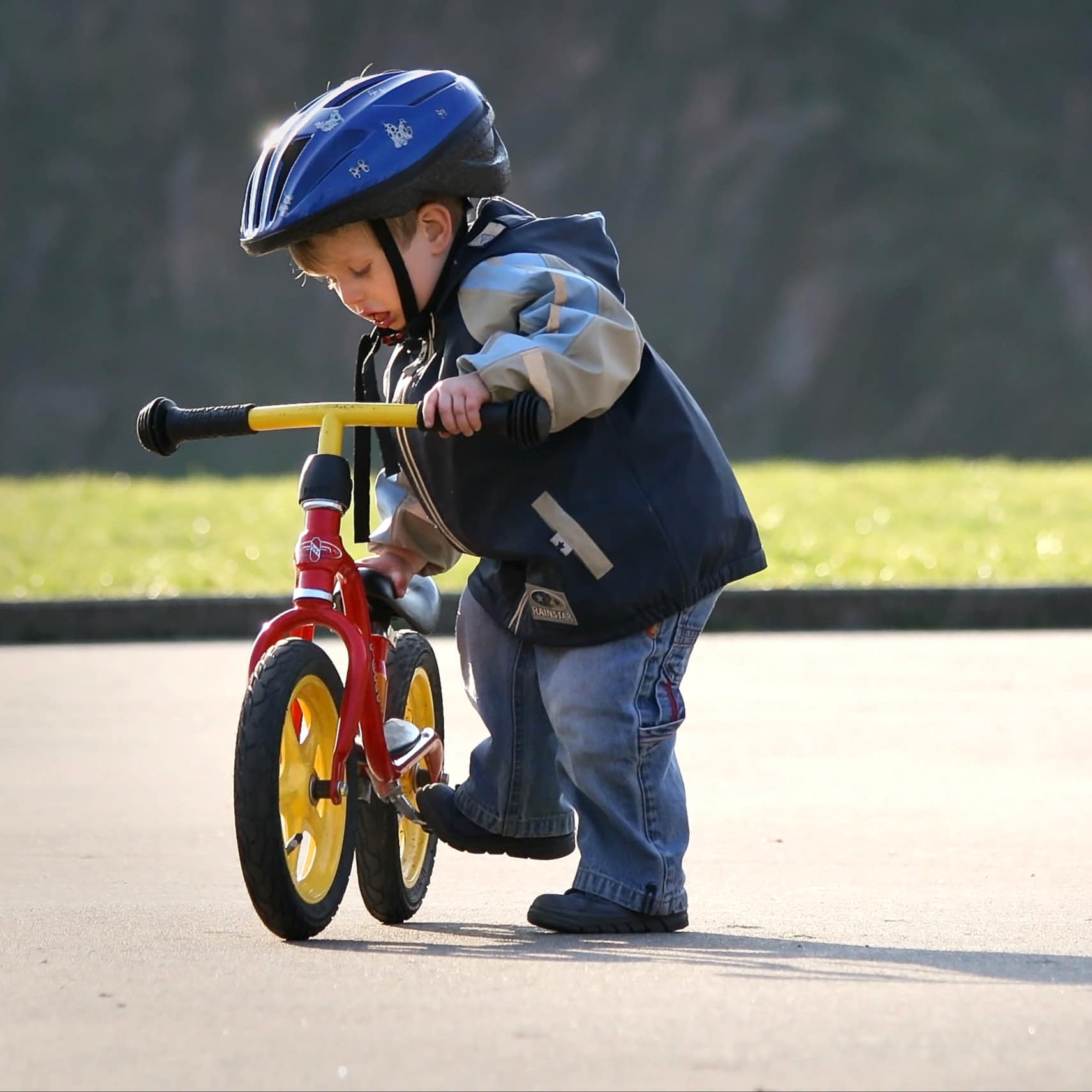 Kleiner Junge in Jeans und Jacke und mit Helm neben seinem Laufrad.