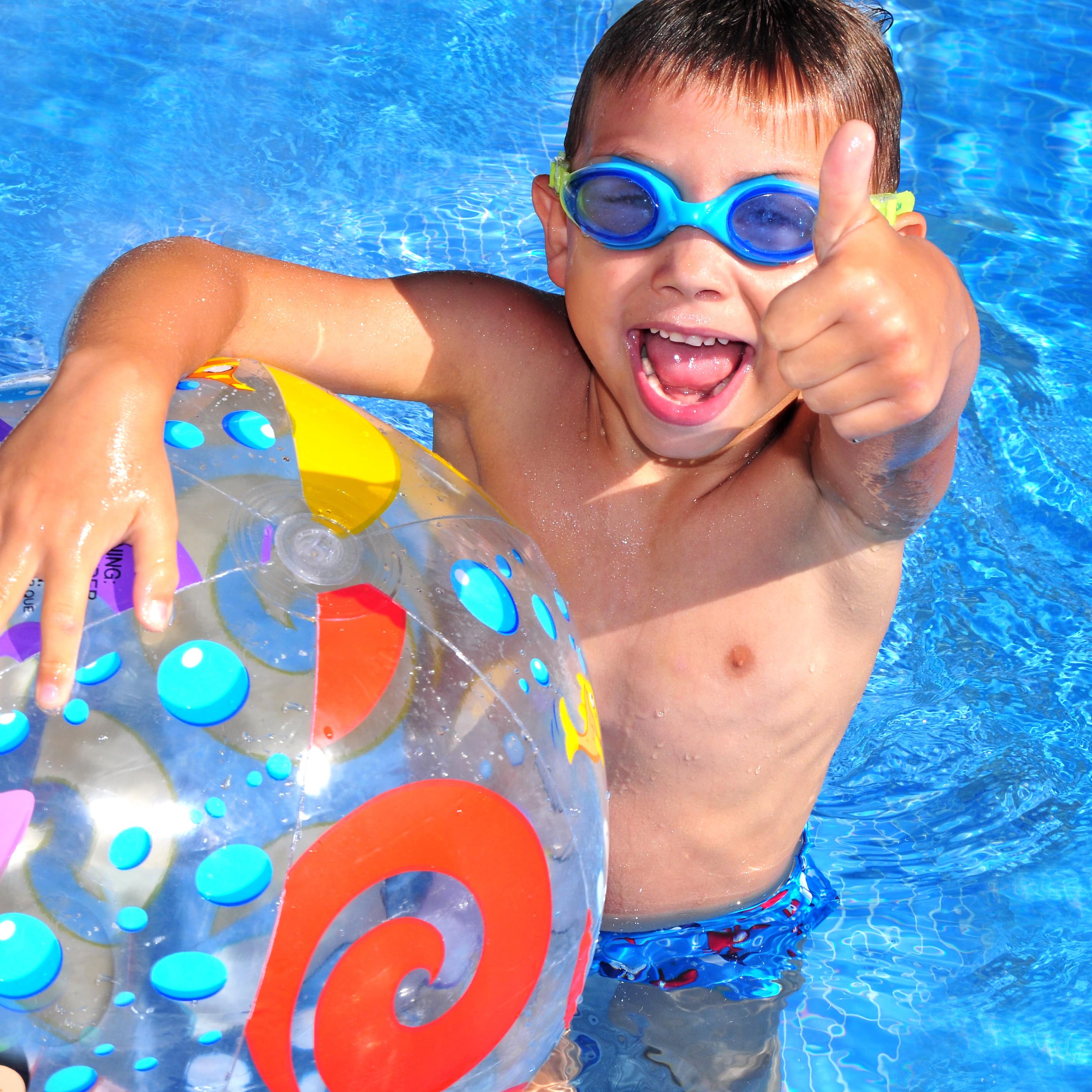 Ein Junge im Schwimmbecken mit Schwimmbrille hält einen Wasserball und streckt einen Daumen nach oben.