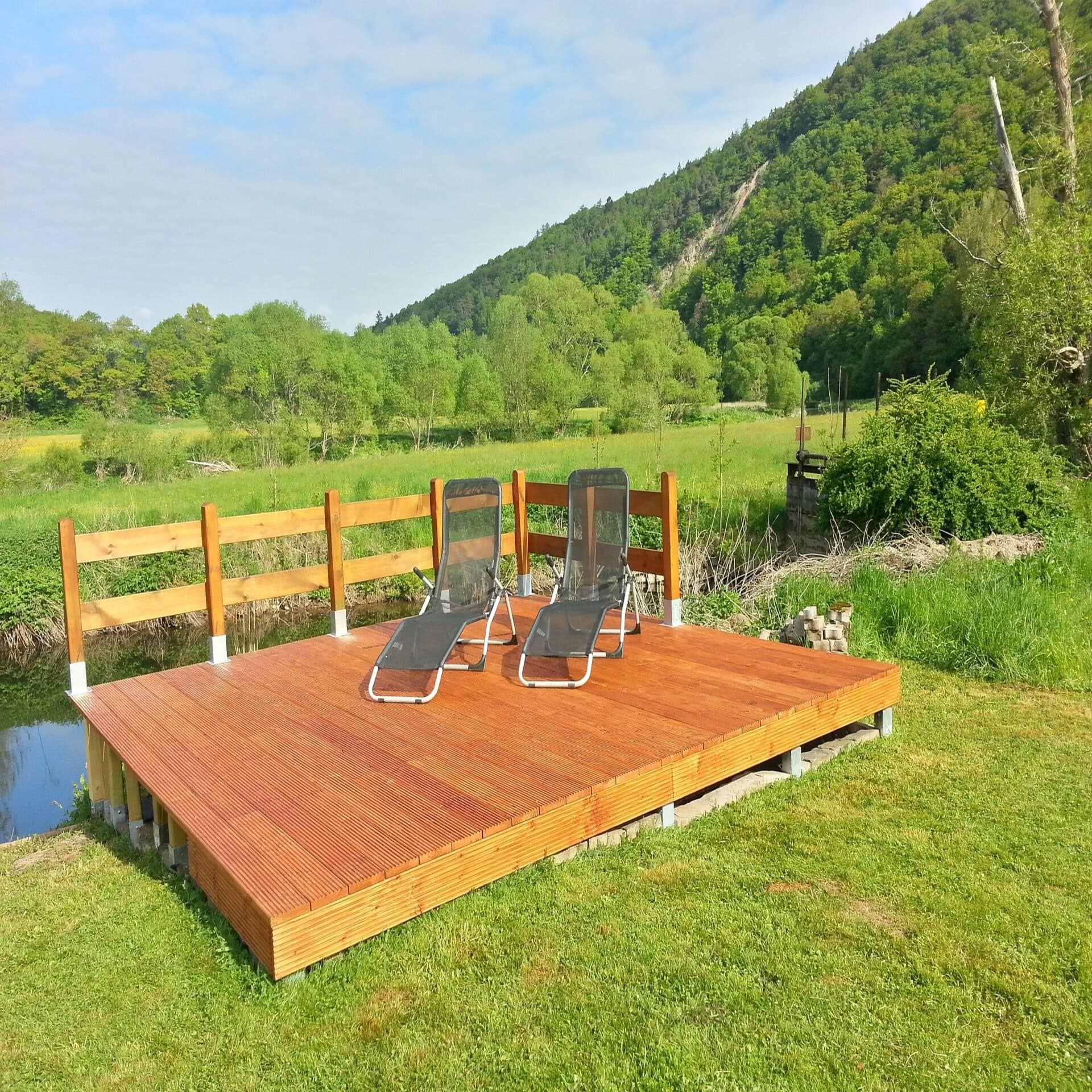 Holzdeck mit 2 Liegestühlen an einem kleinen Wasserlauf, dahinter die grüne Natur. Die Sonne scheint. 