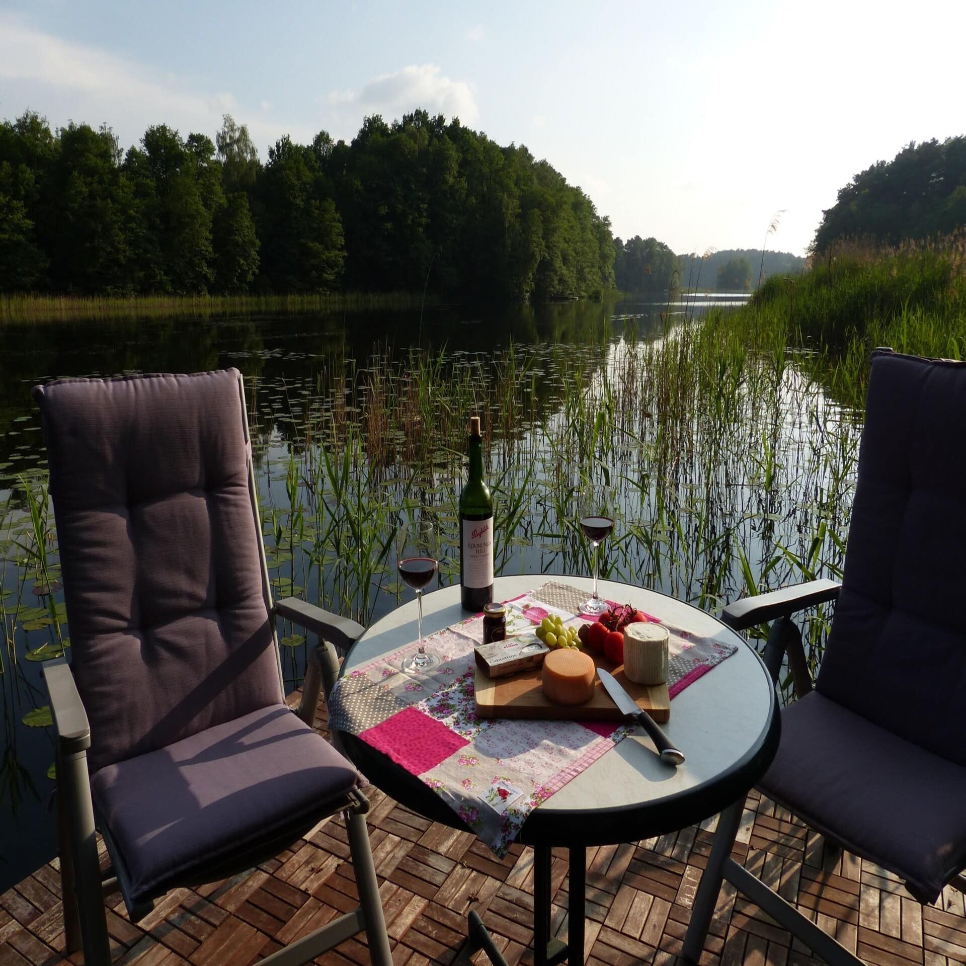 Terrasse direkt am Wasser, darauf 2 Stühle und ein Tisch mit Käseplatte und Rotwein.