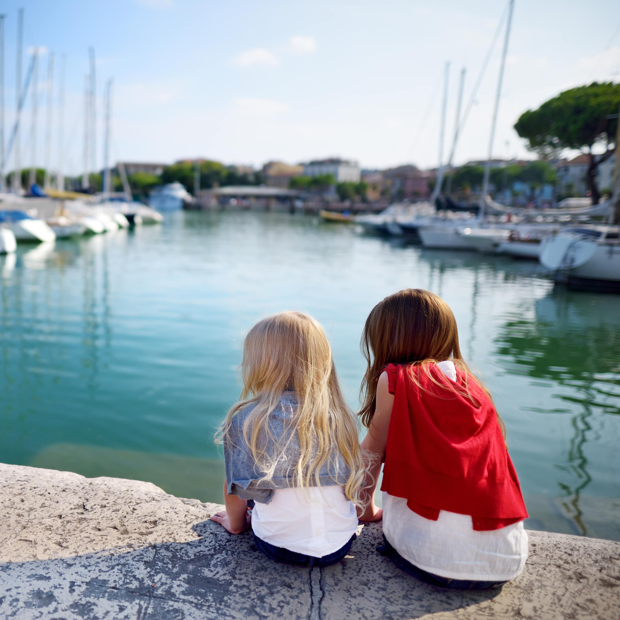 Rückenansicht: 2 Mädchen sitzen auf einer Mauer am Hafen und schauen auf die Boote.