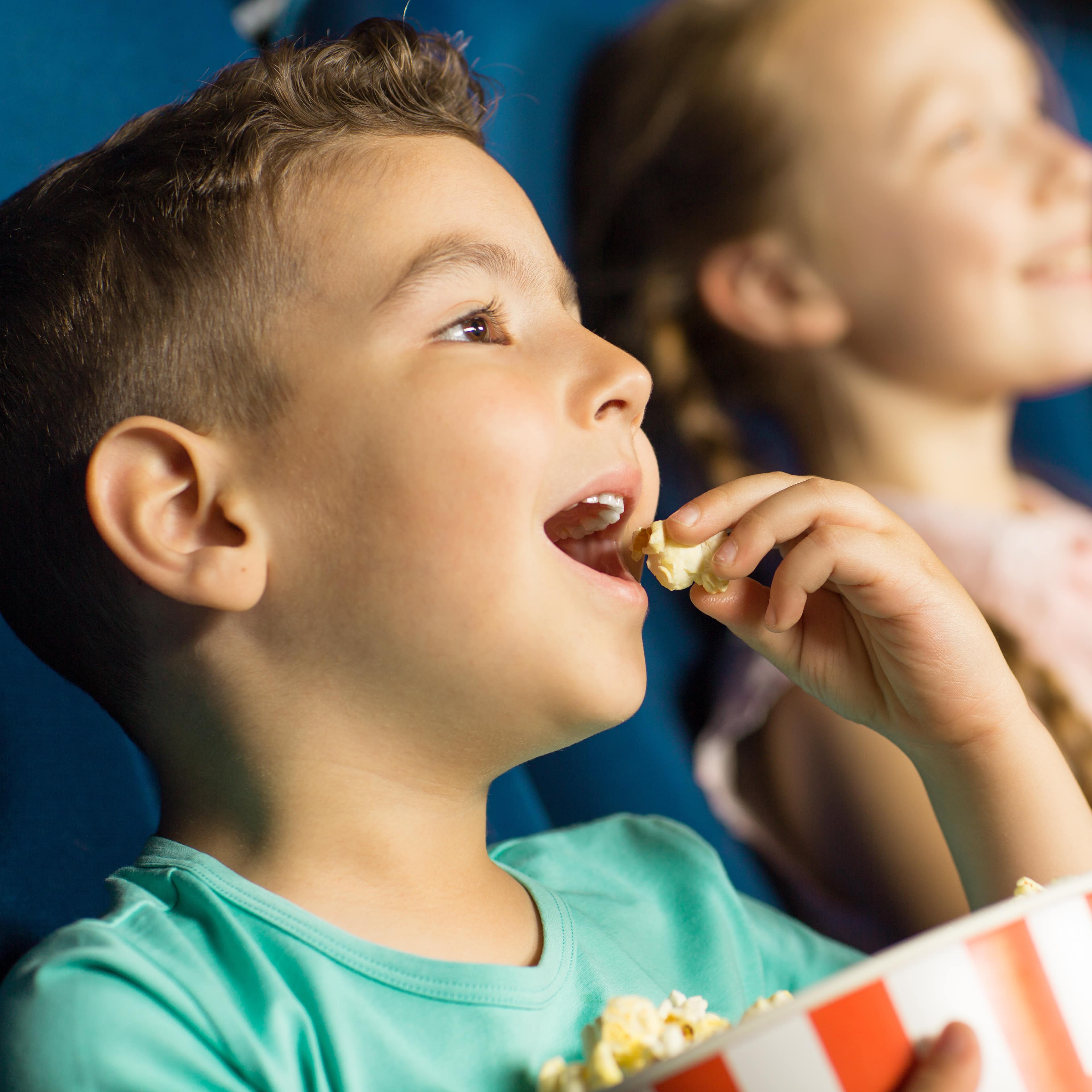 Ein Junge sitzt in einem blauen Kinositz und isst Popcorn aus einen rot-weiß gestreiften Eimer.