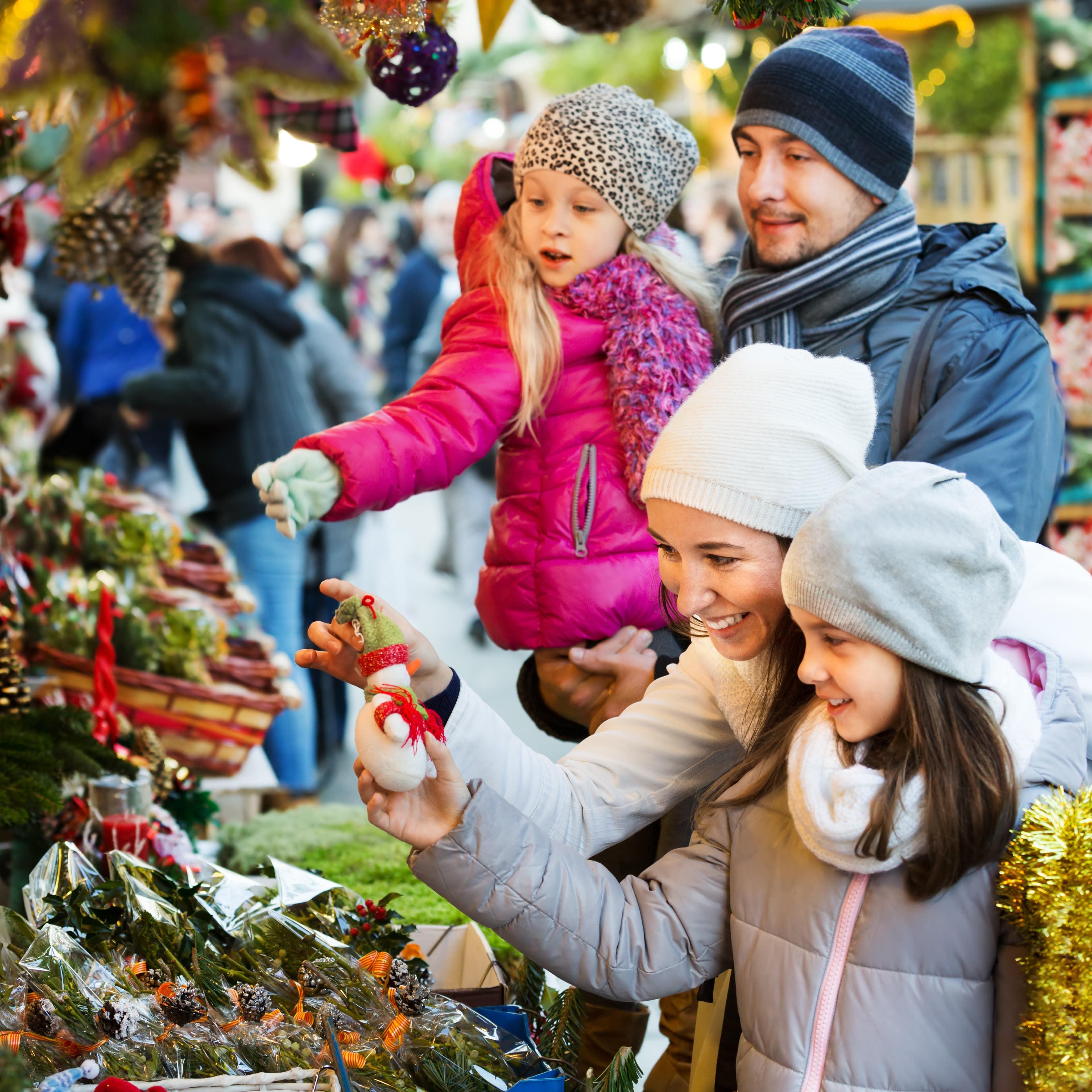 Familie mit 2 Töchtern schauen sich weihnachtliche Dekoration an einem Stand auf dem Weihnachtsmarkt an.
