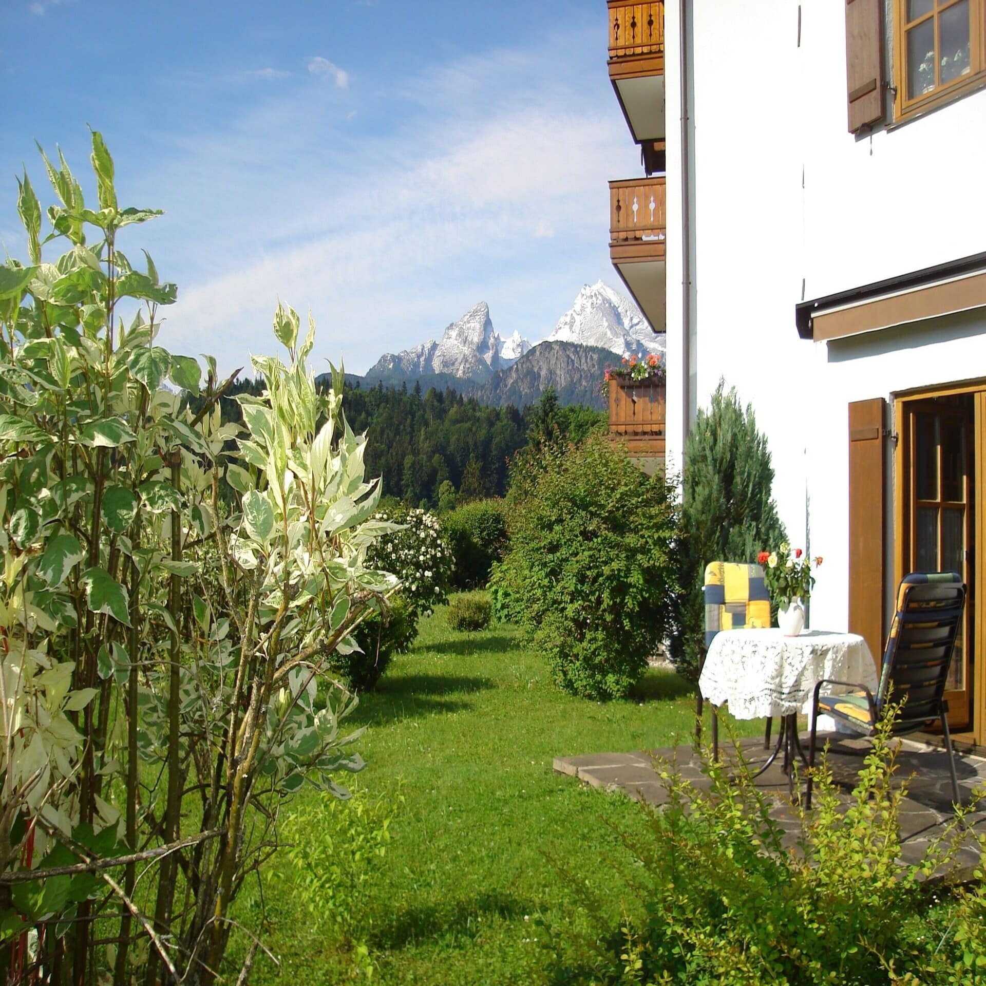 Ecke eines Hauses mit Terrasse und Balkonen. Blick durch den grünen Garten auf die Berge.