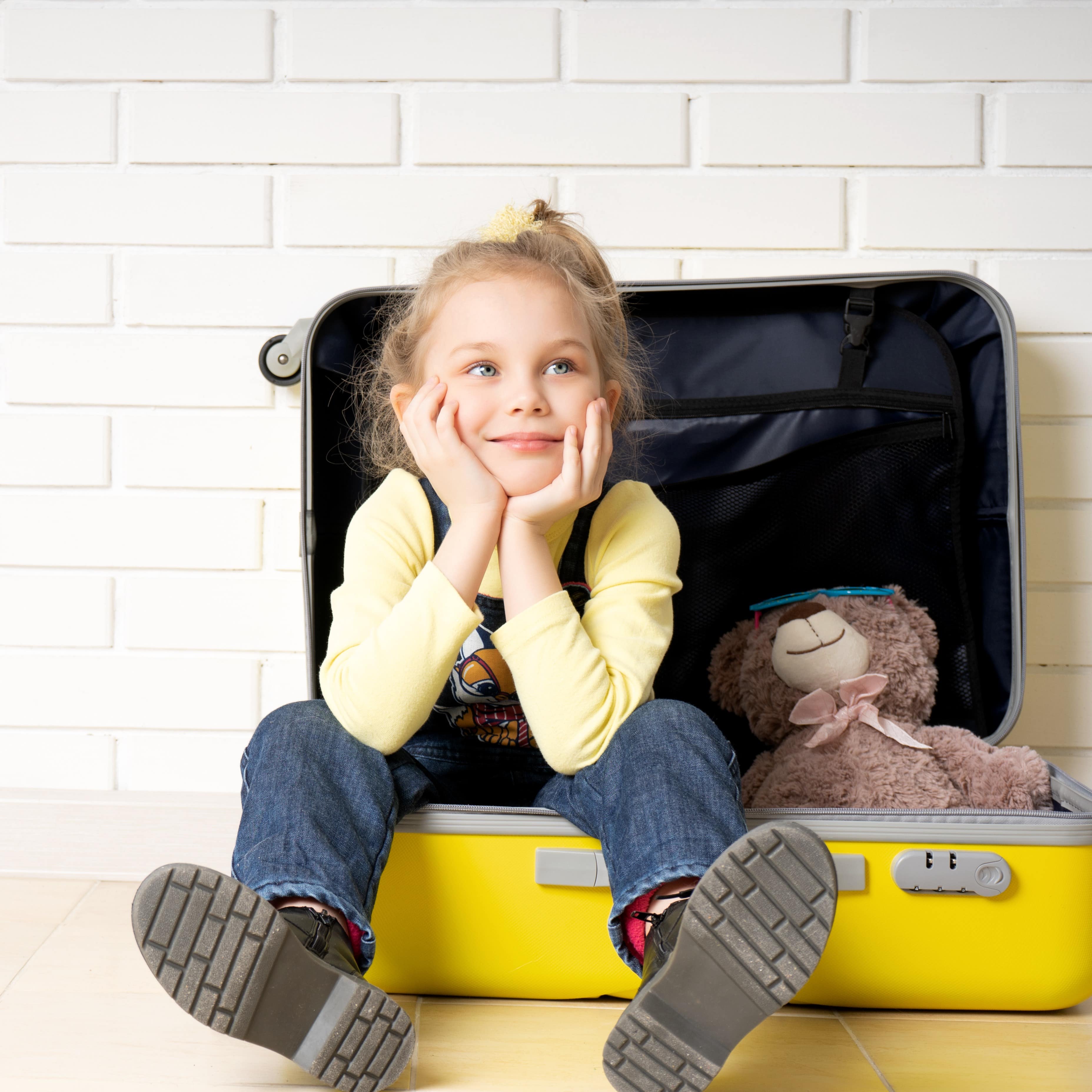 Ein Mädchen in Jeans und gelbem Pulli sitzt mit einem Teddy in einem geöffneten gelben Koffer.