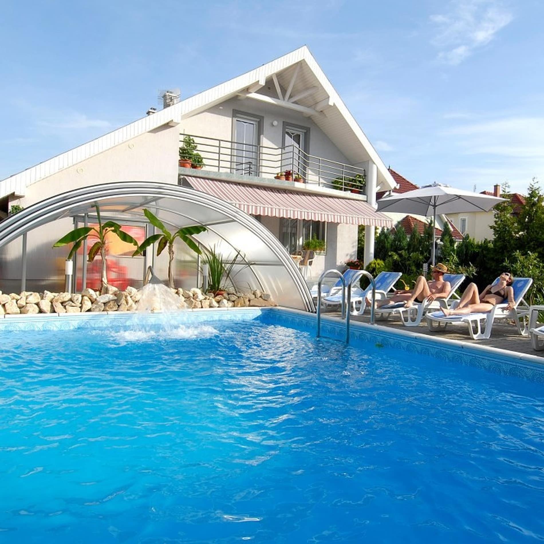 Großer Pool mit sprudelndem Wassereinlauf vor einem weißen Haus mit zwei Ferienwohnungen