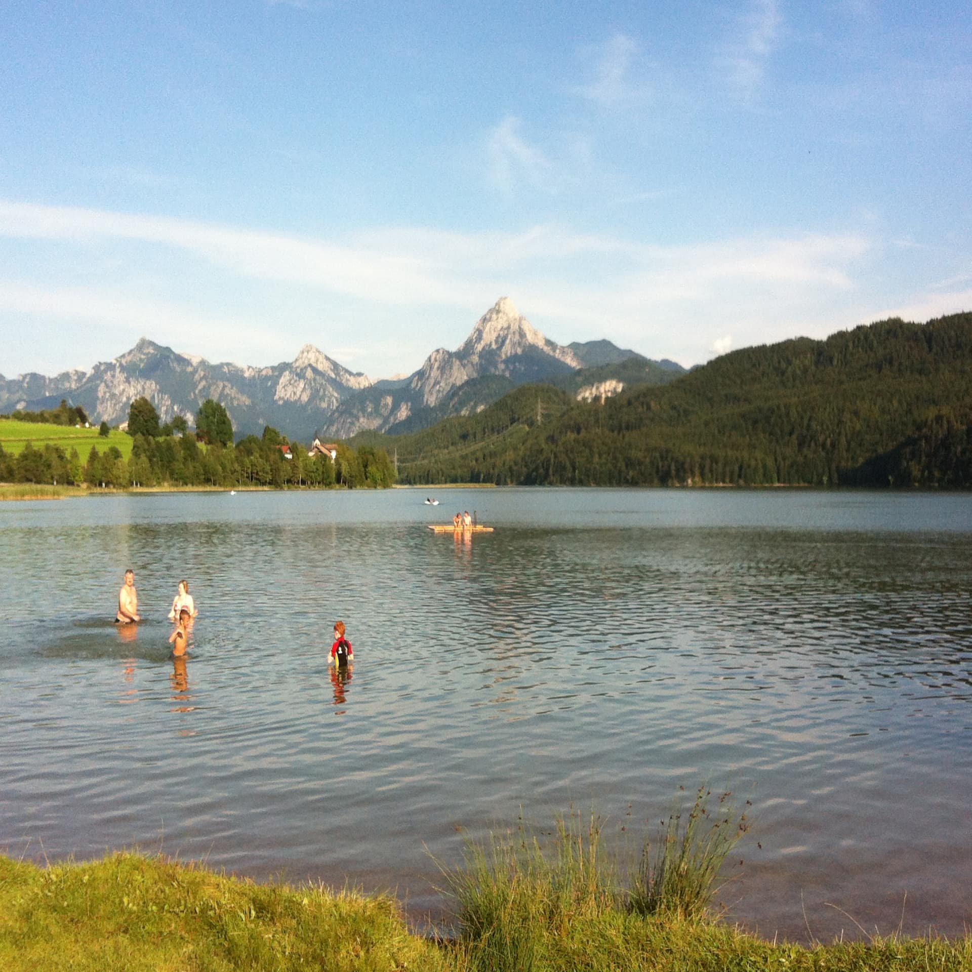 Kinder und Erwachsene plantschen in einem See. Im Hintergrund Wälder und Berge. 