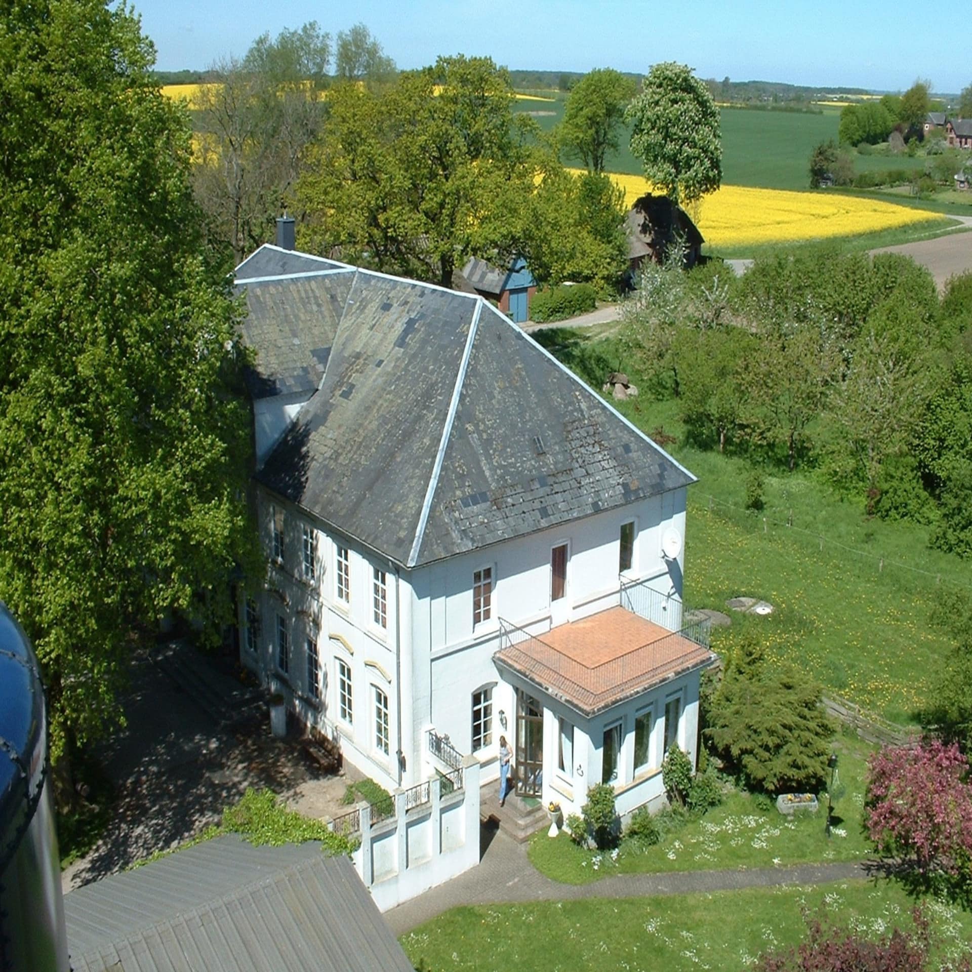 Panoramablick von oben auf ein weißes Bauernhaus inmitten grüner und gelber Felder. 