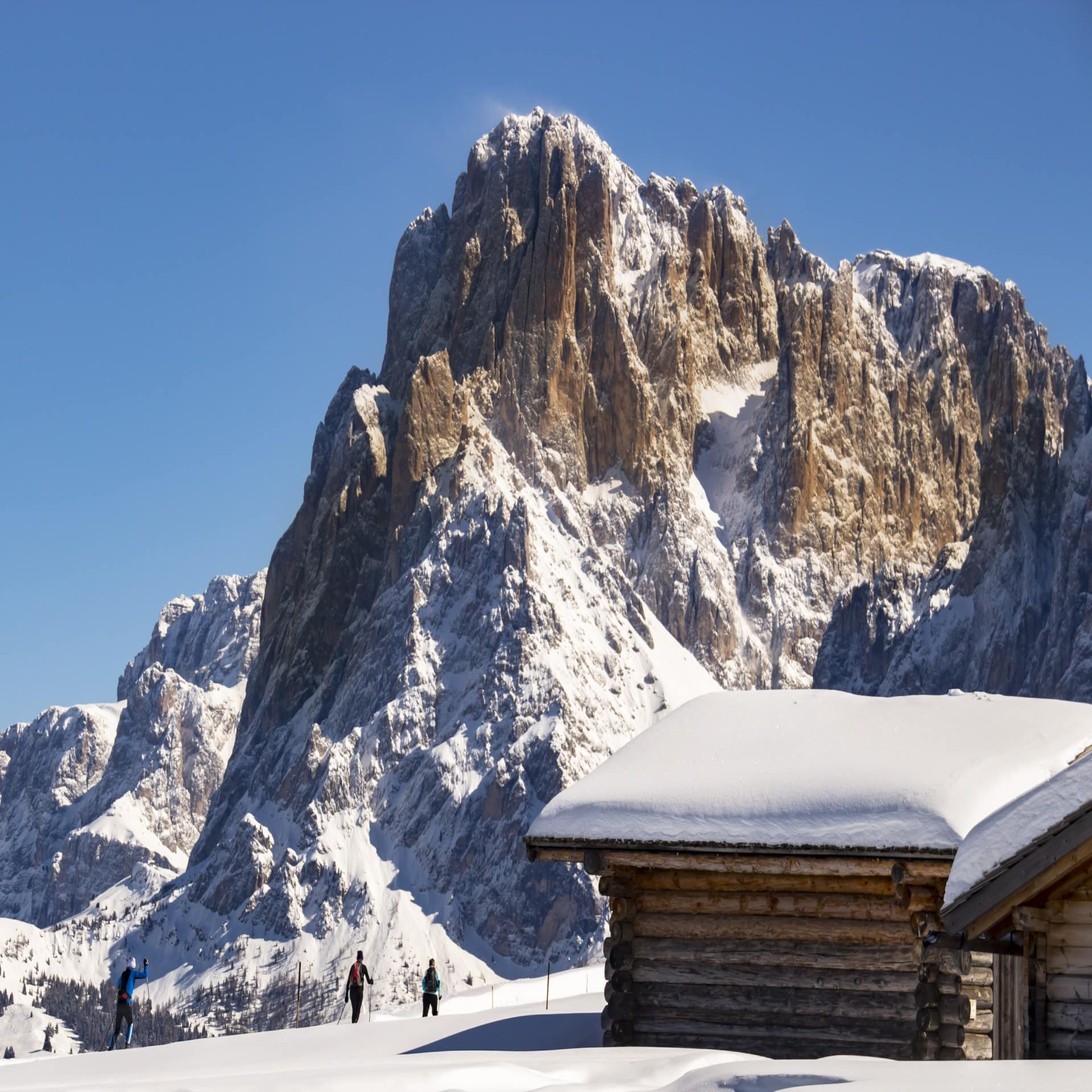 3 Langläufer steuern auf eine verschneite Hütte zu, ein Bergmassiv im Hintergrund. 