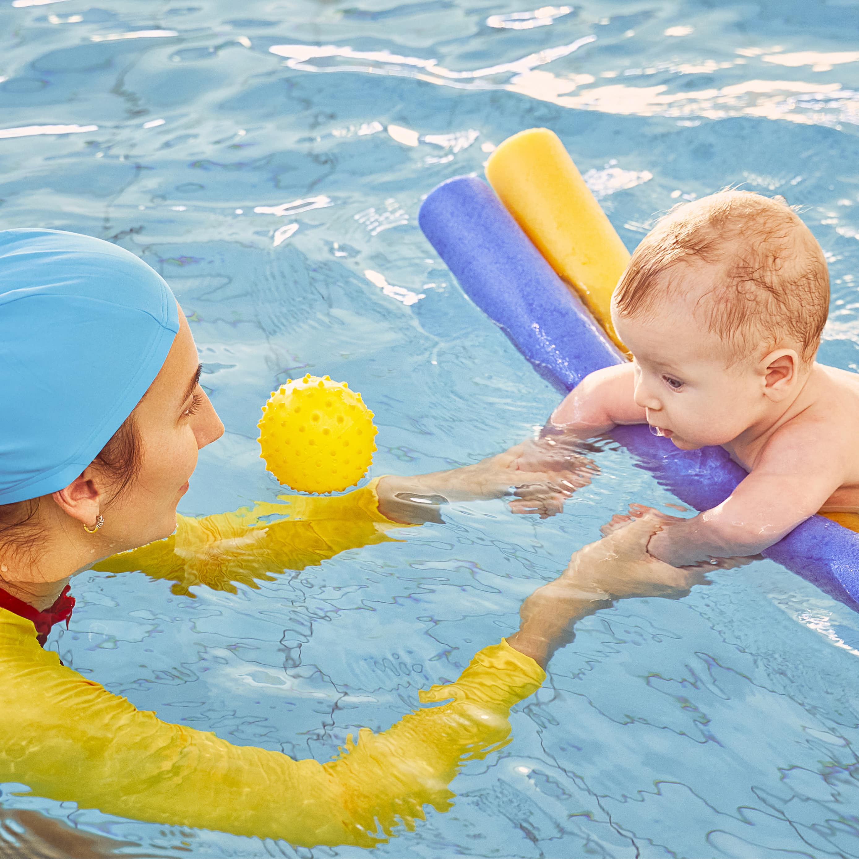 Babyschwimmen: Frau in langem Schwimmoutfit und mit Badekappe zieht ein Baby auf Schwimmnu-deln durchs Wasser.