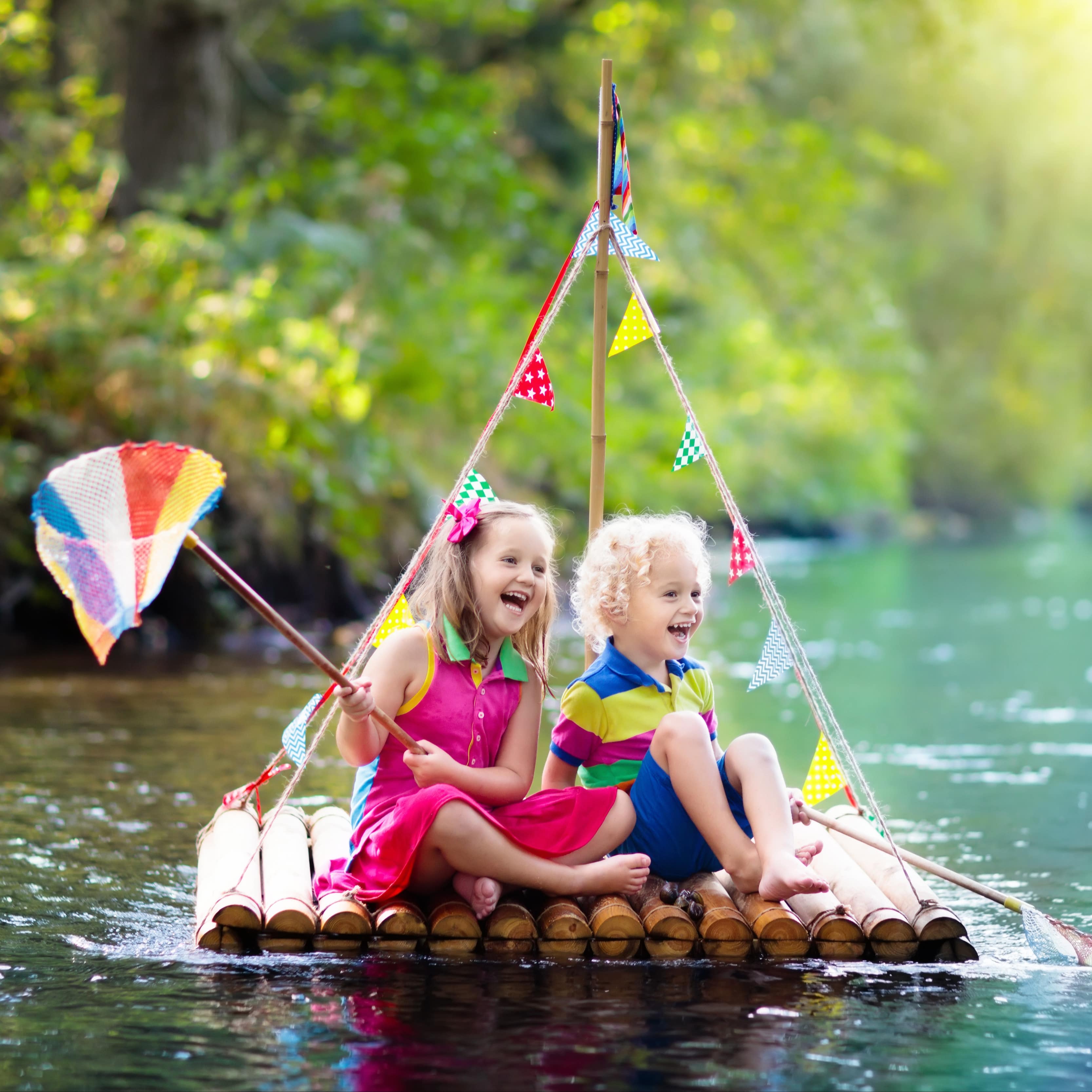 2 Kinder in bunter Sommerkleidung auf einem kleinen Holzfloß mit bunten Wimpeln. 