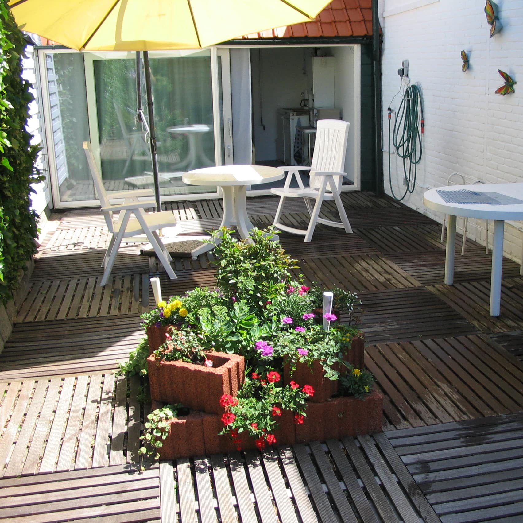Ferienwohnung in den Niederlanden mit großer Sonnenterrasse, darauf Blumen und Esstisch mit Stühlen unter einem Sonnenschirm