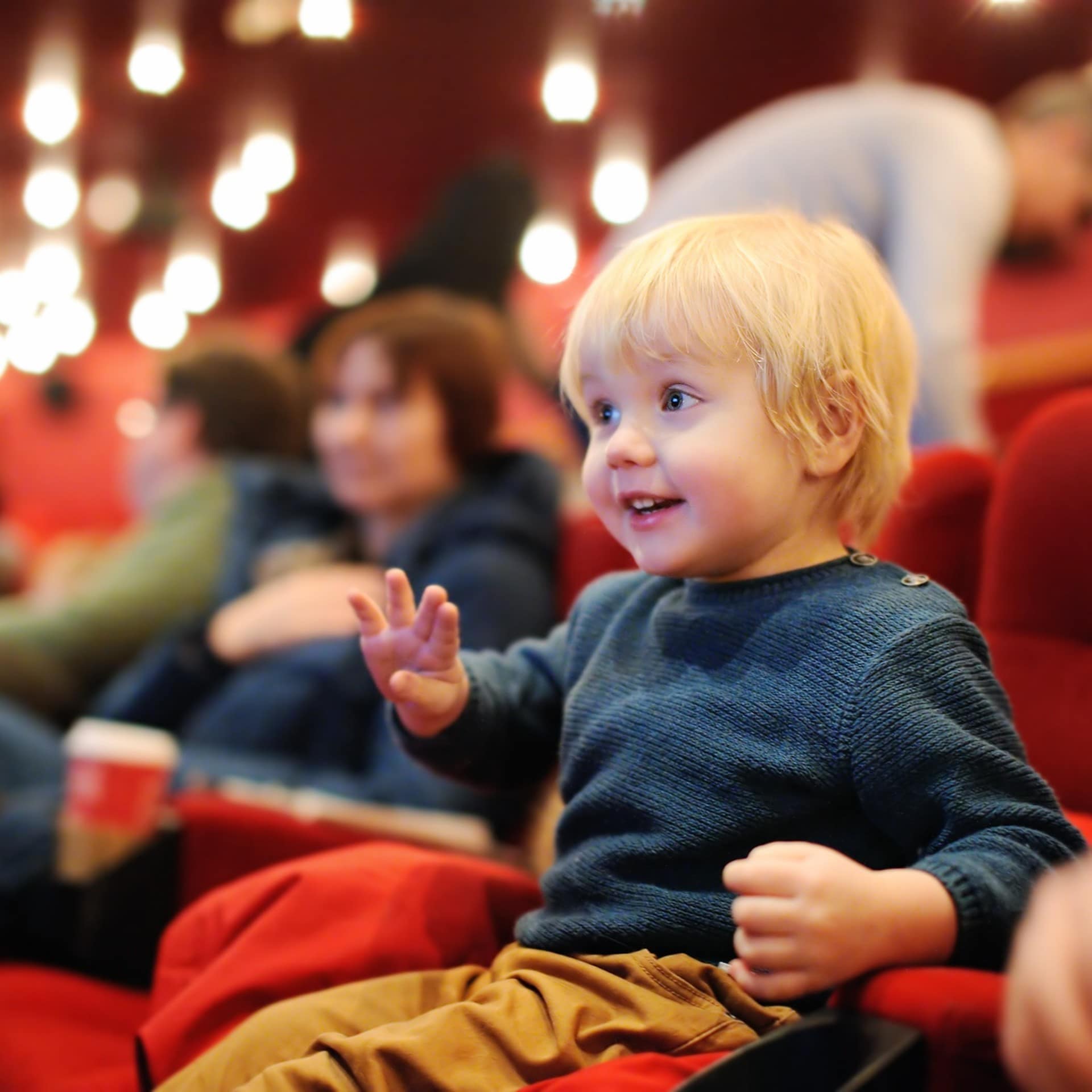 Blondes Kleinkind mit blauem Pulli auf einem roten Sitz im Kino schaut auf die Leinwand.