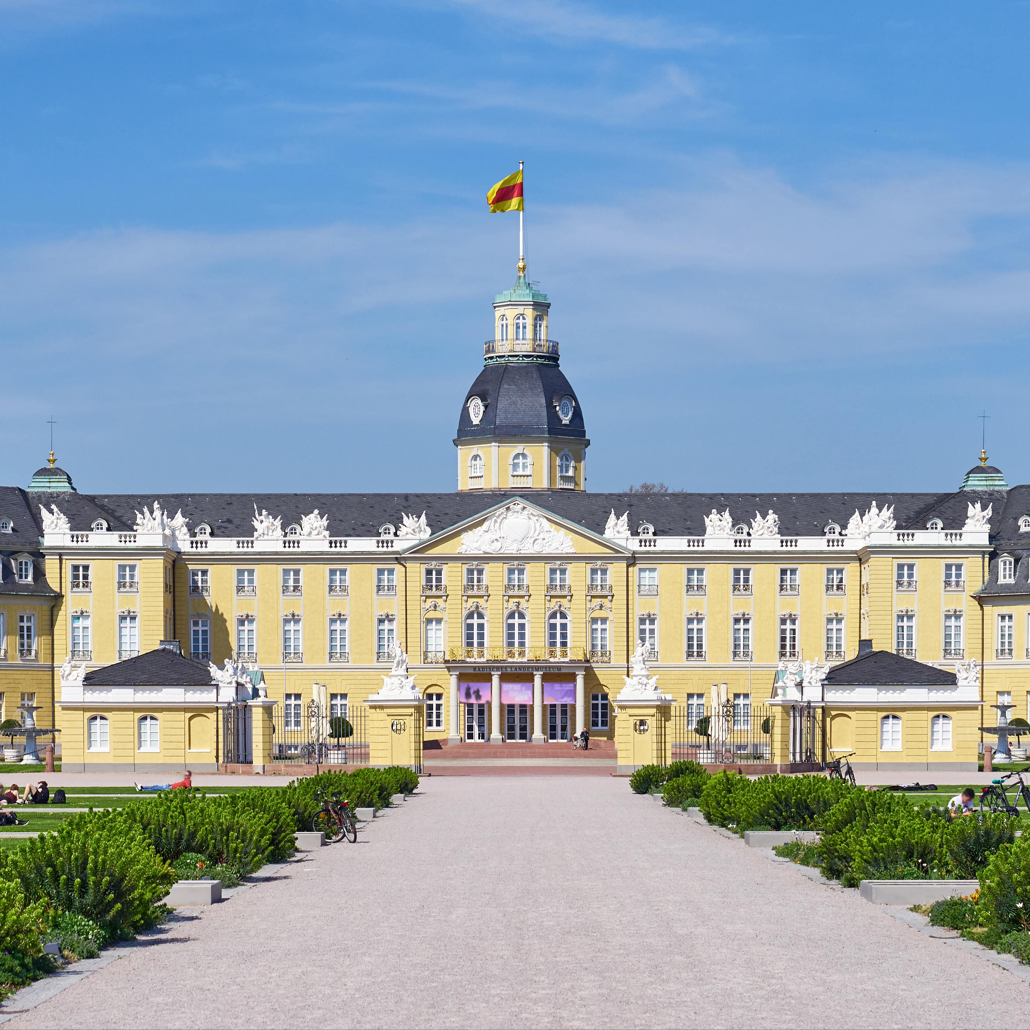 Blick auf das Schloss in Karlsruhe. Auf den Wiesen davor sitzen Leute in der Sonne.