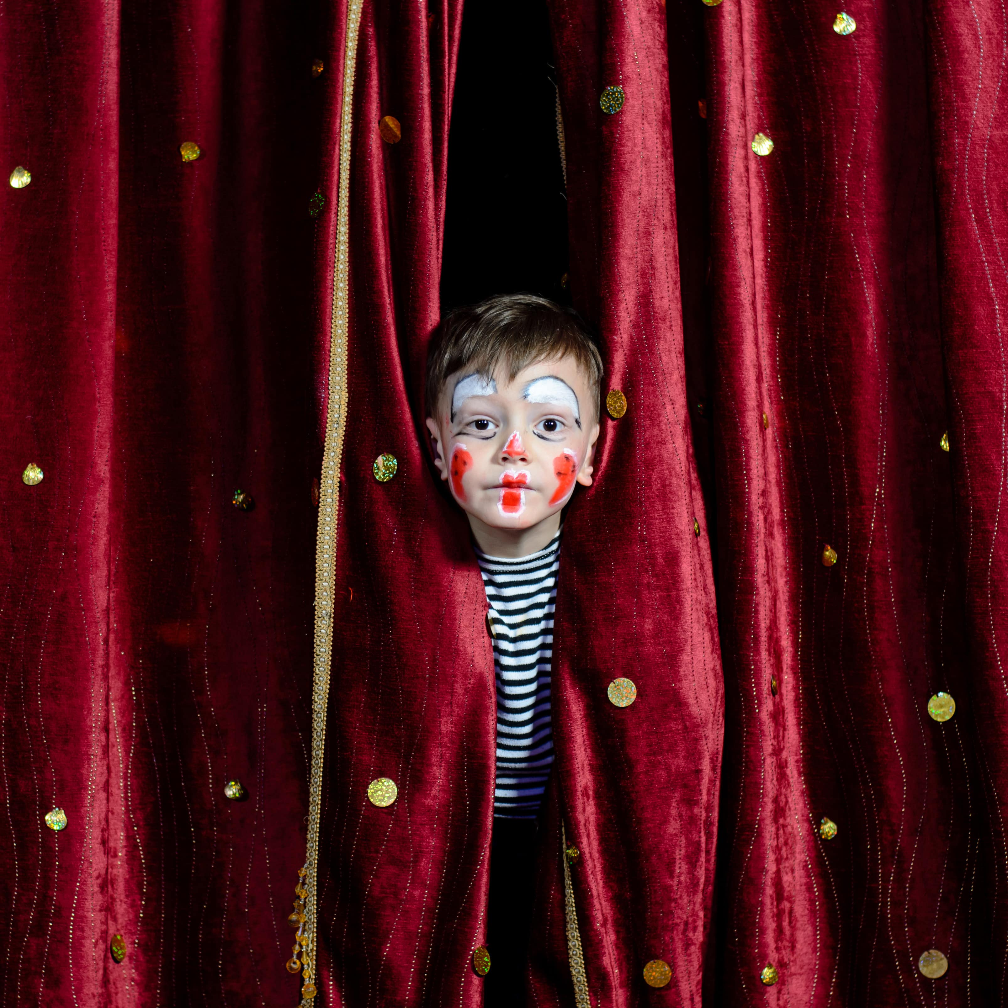 Ein Junge mit geschminktem Clownsgesicht steckt den Kopf durch einen dunkelroten Vorhang mit goldenen Verzierungen.
