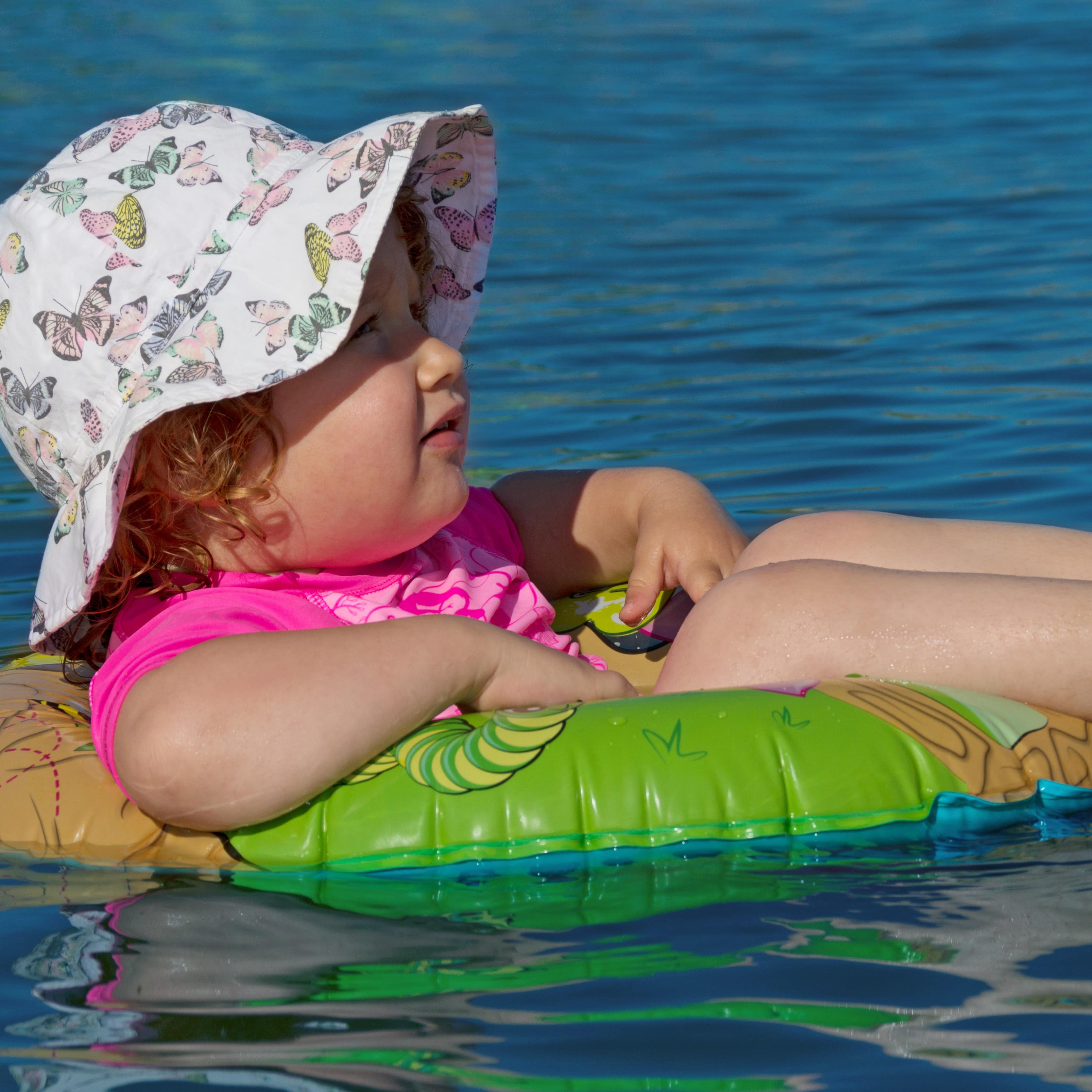 Kleines Mädchen in pinkem Badeanzug und mit Hut sitzt in einem Schwimmring im Wasser.
