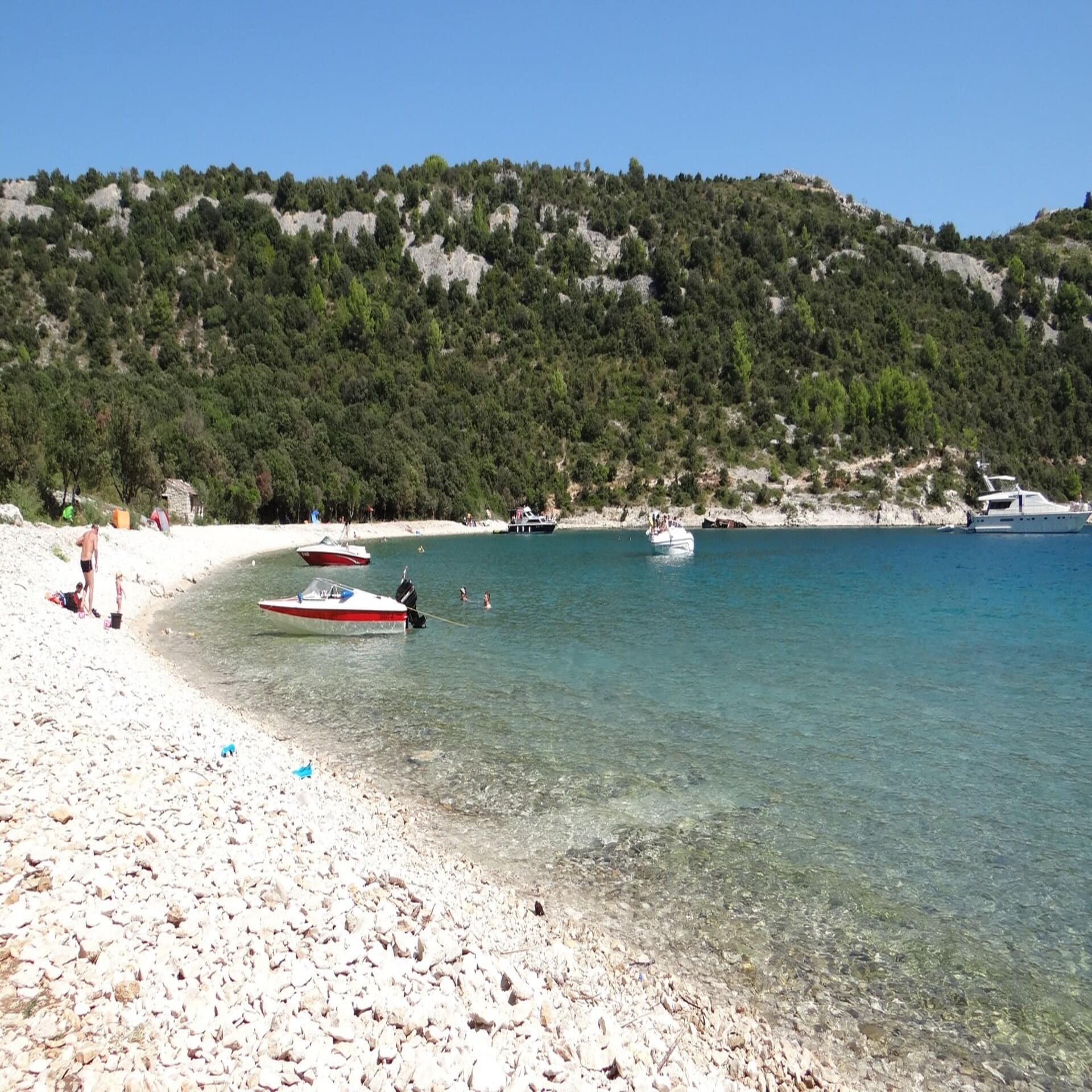 Personen am Kiesstrand, im Wasser einige Boote. Die Sonne scheint. 