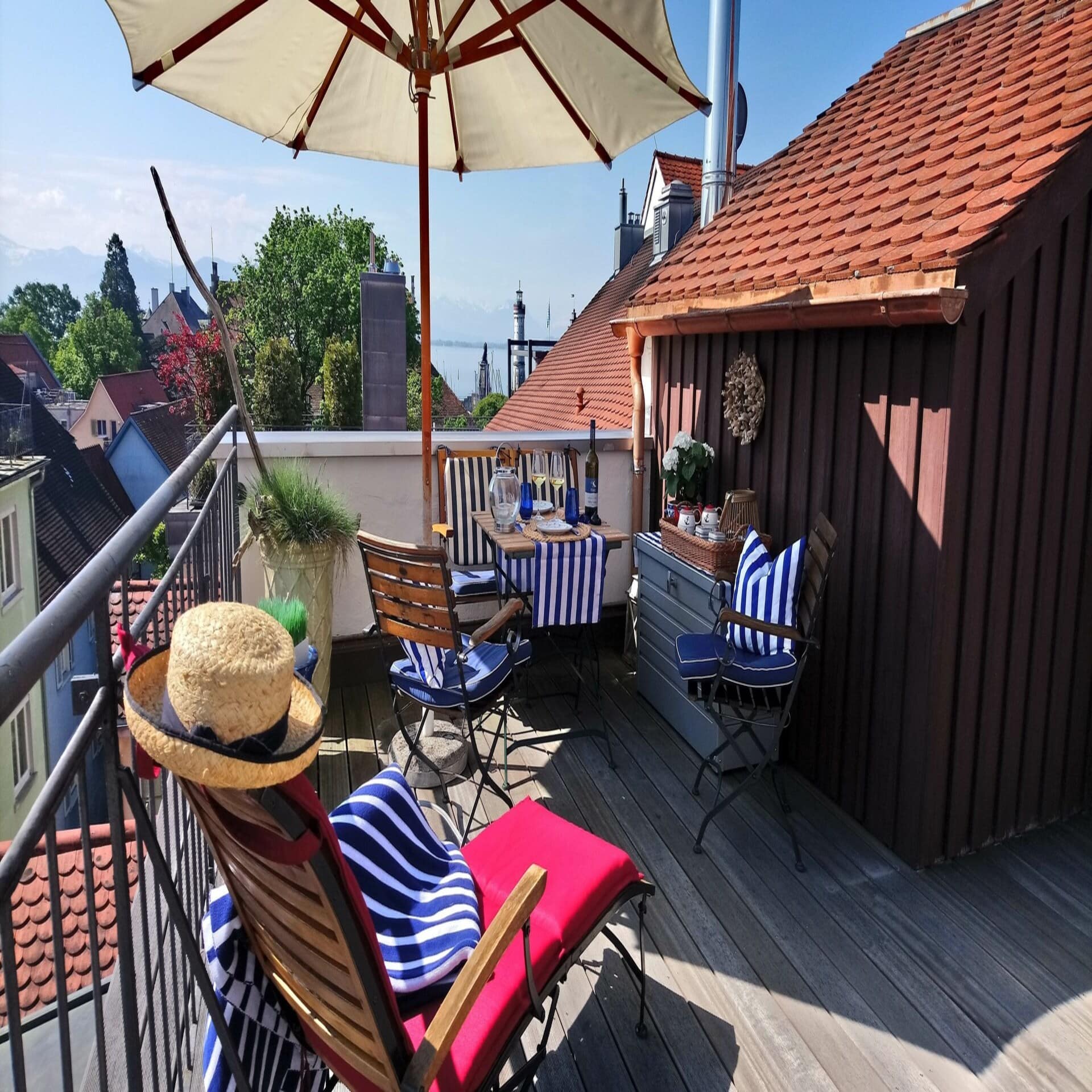 Dachterrasse mit Tisch, Stühlen, Liegestuhl und Sonnenschirm. Blick über die Häuserdächer von Lindau und auf den Bodensee. 