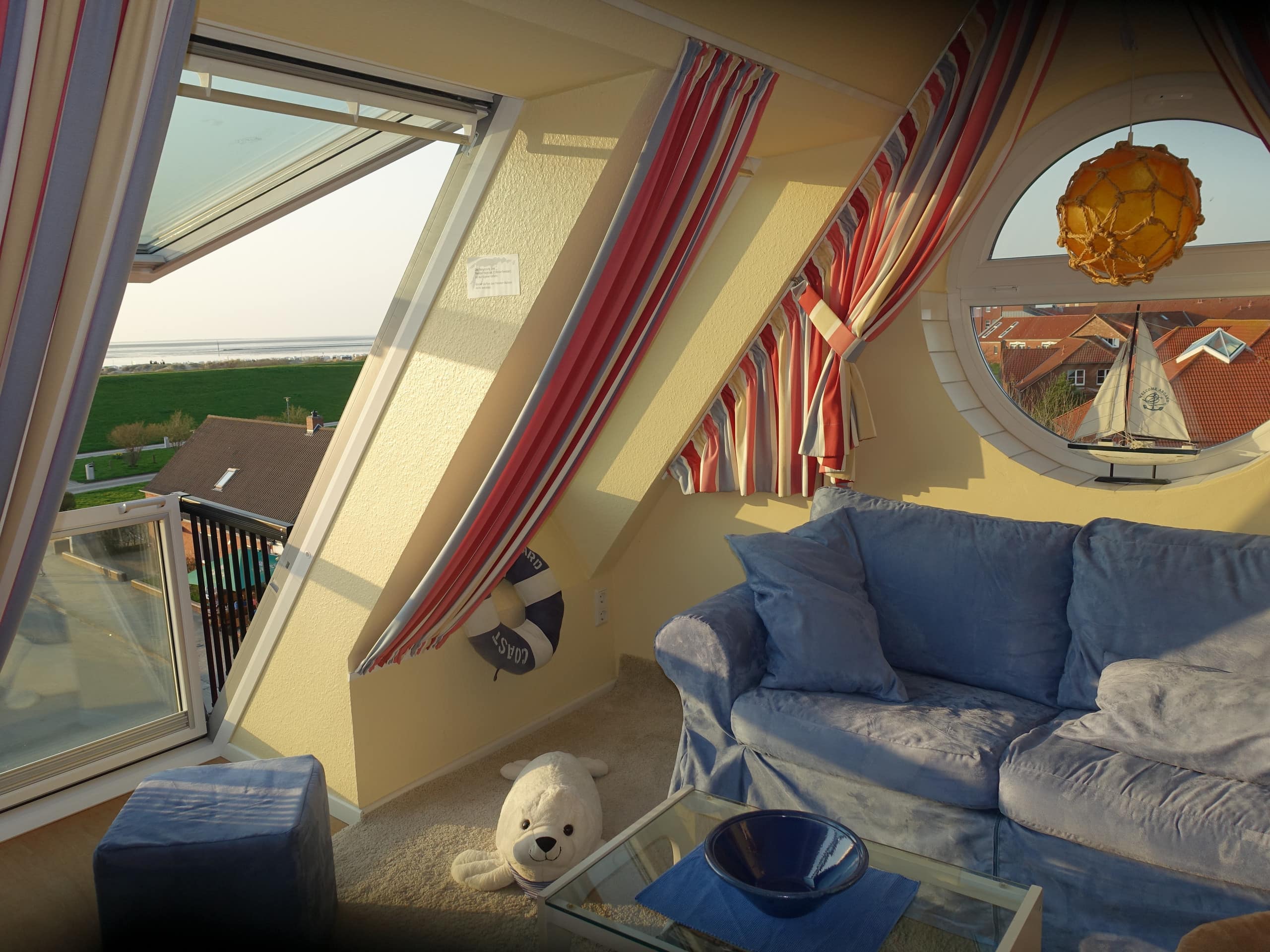 Wohnzimmer einer Ferienwohnung in Norddeich im Dachgeschoss, durch das geöffnete Fenster ist das Meer zu sehen.