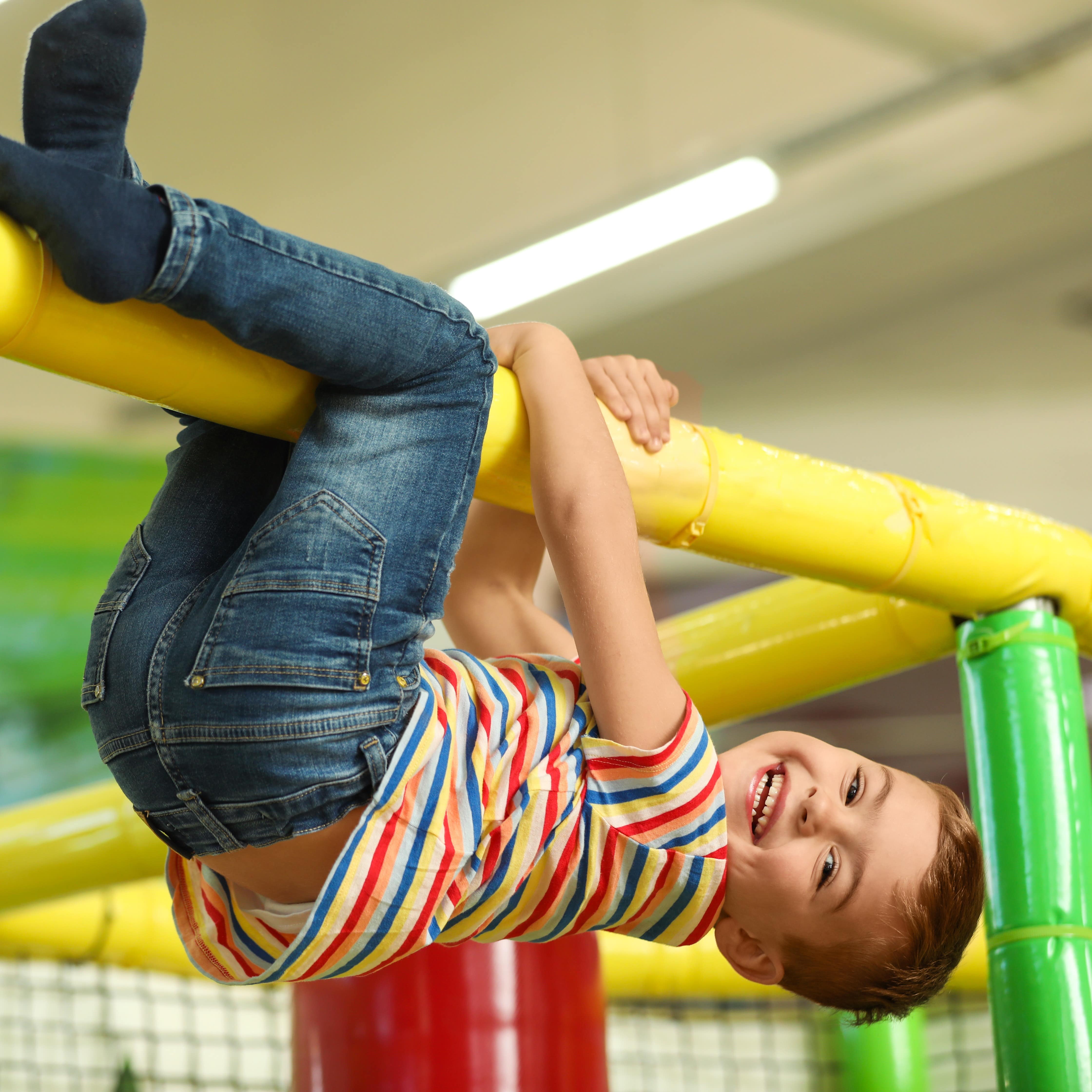 Junge in Jeans und buntem T-Shirt hängt an einem Klettergerüst im Indoor-Spielplatz.