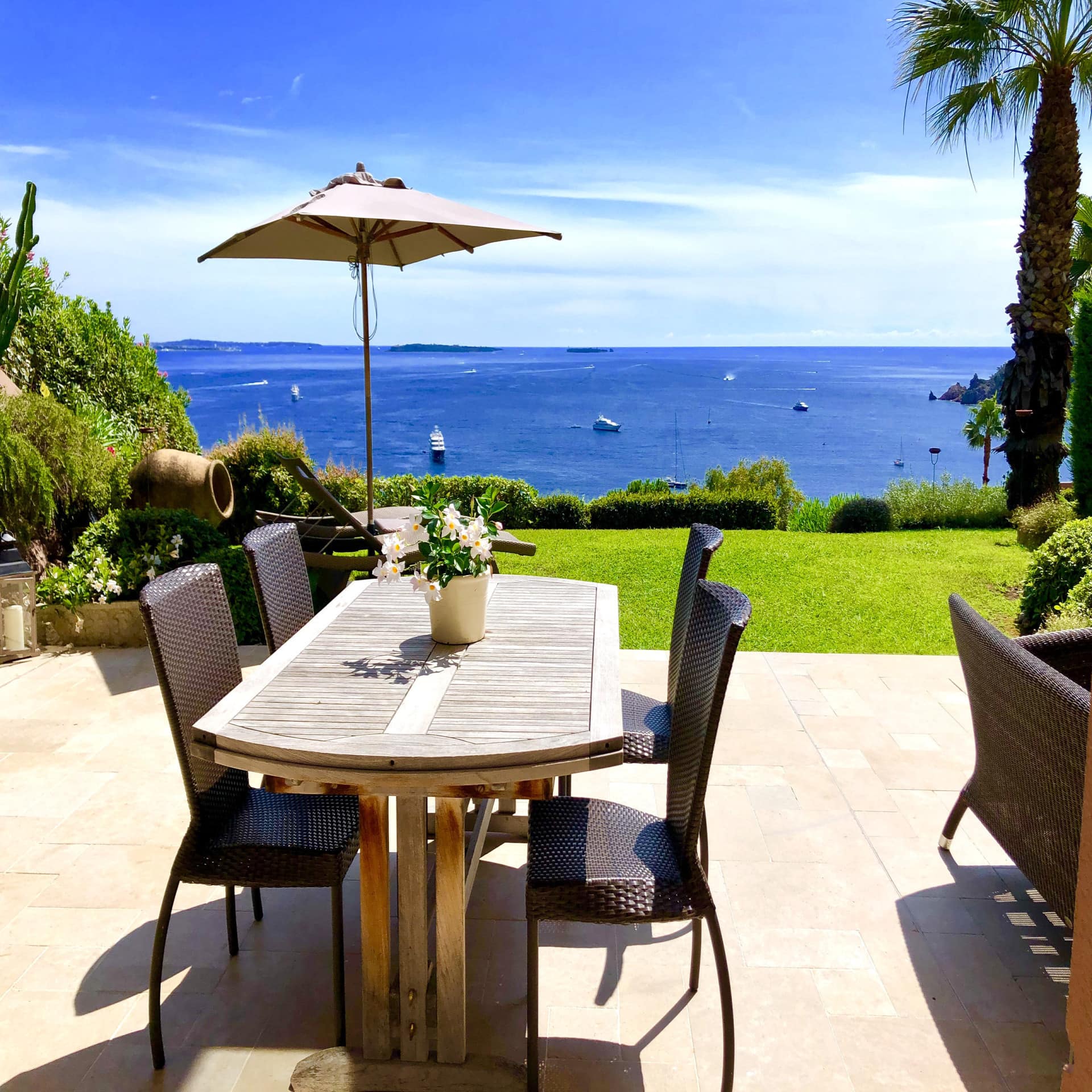 Terrasse mit Tisch, 4 Stühlen und Sonnenschirm. Blick über einen manikürten Rasen auf das Meer und einige Boote.