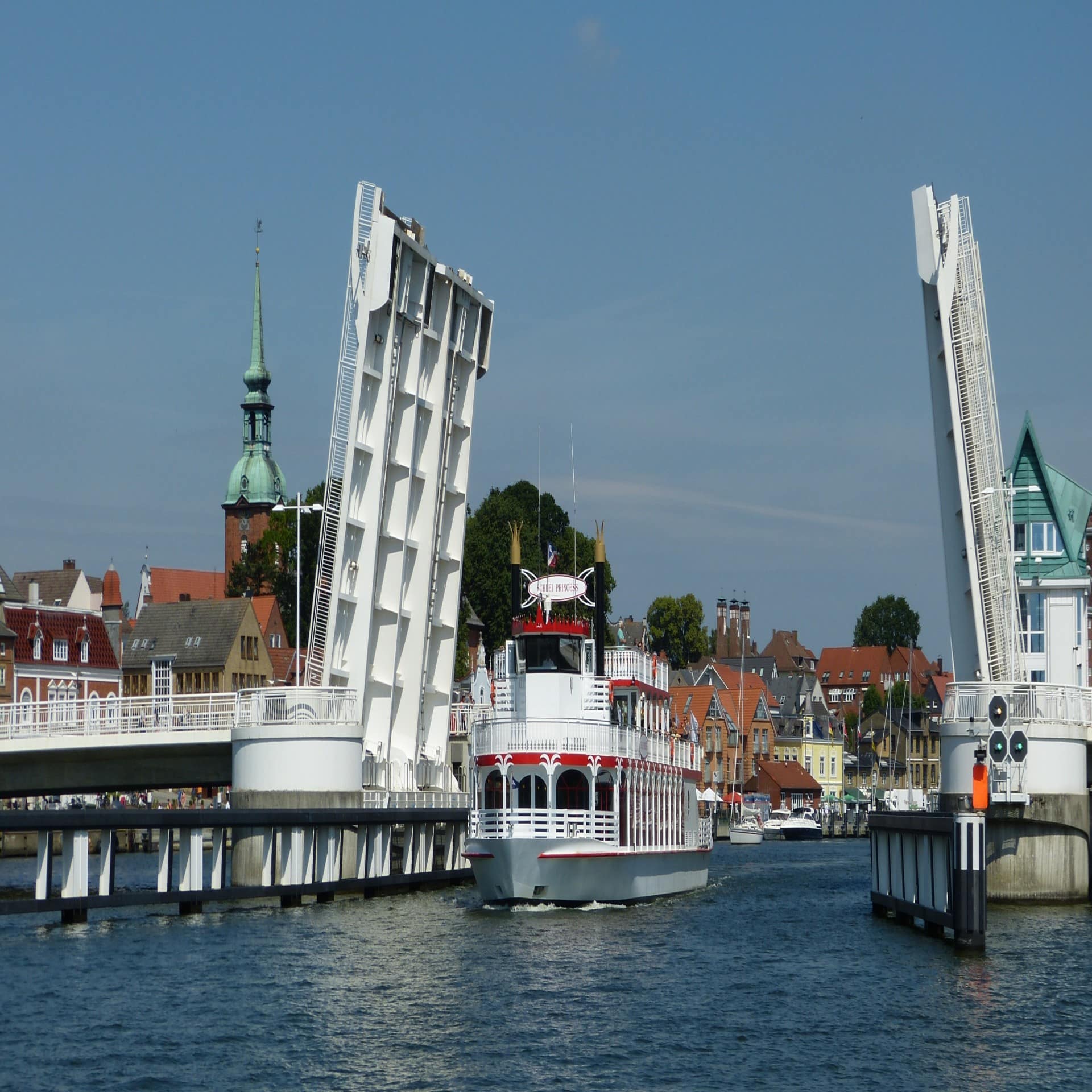 Ein weißes Schiff fährt unter der geöffneten Klappbrücke durch, im Hintergrund Hafen und Häuser von Kappeln.