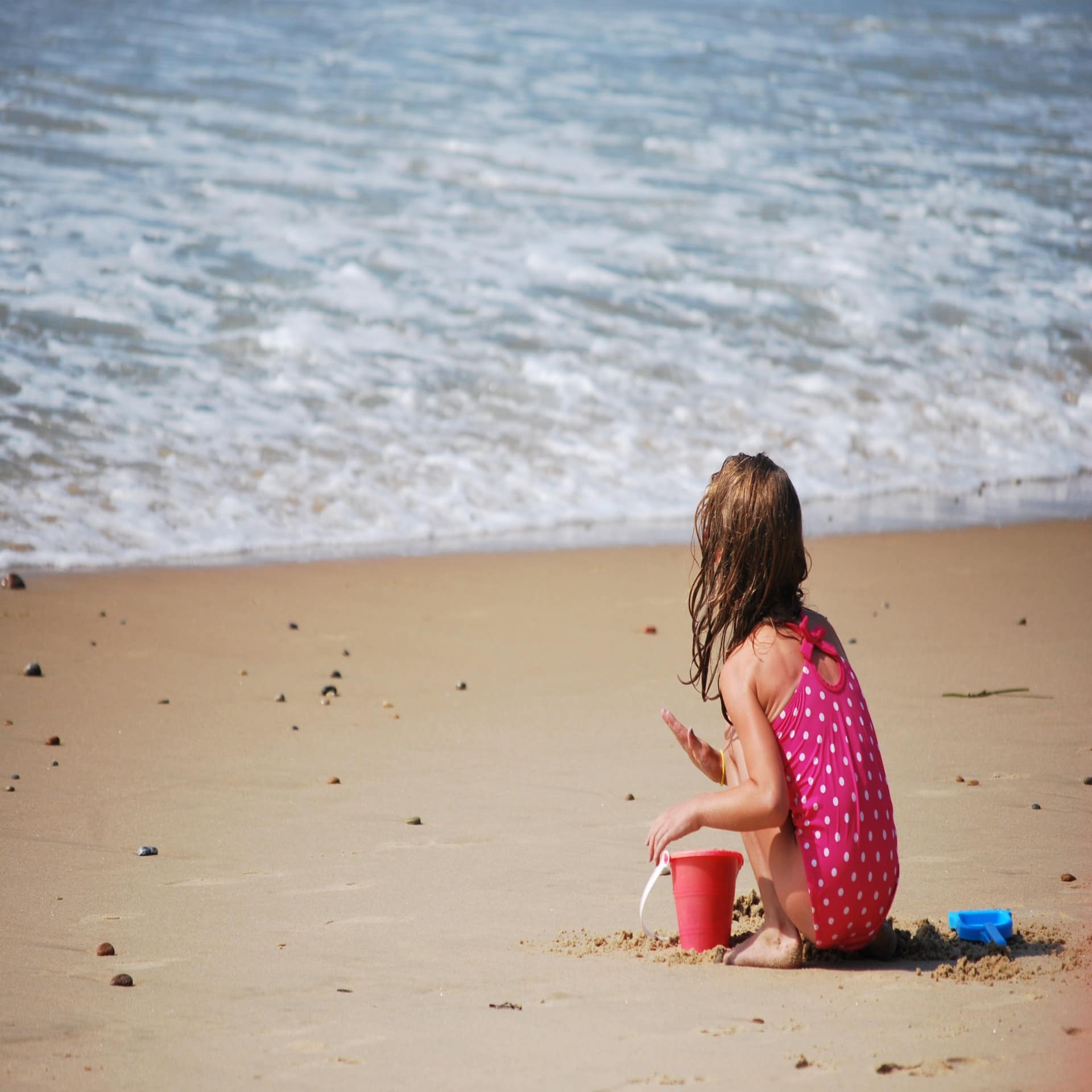 Mädchen in pinkem Badeanzug spielt mit Eimer und Schaufel am Strand. Das Meer ist seicht. 
